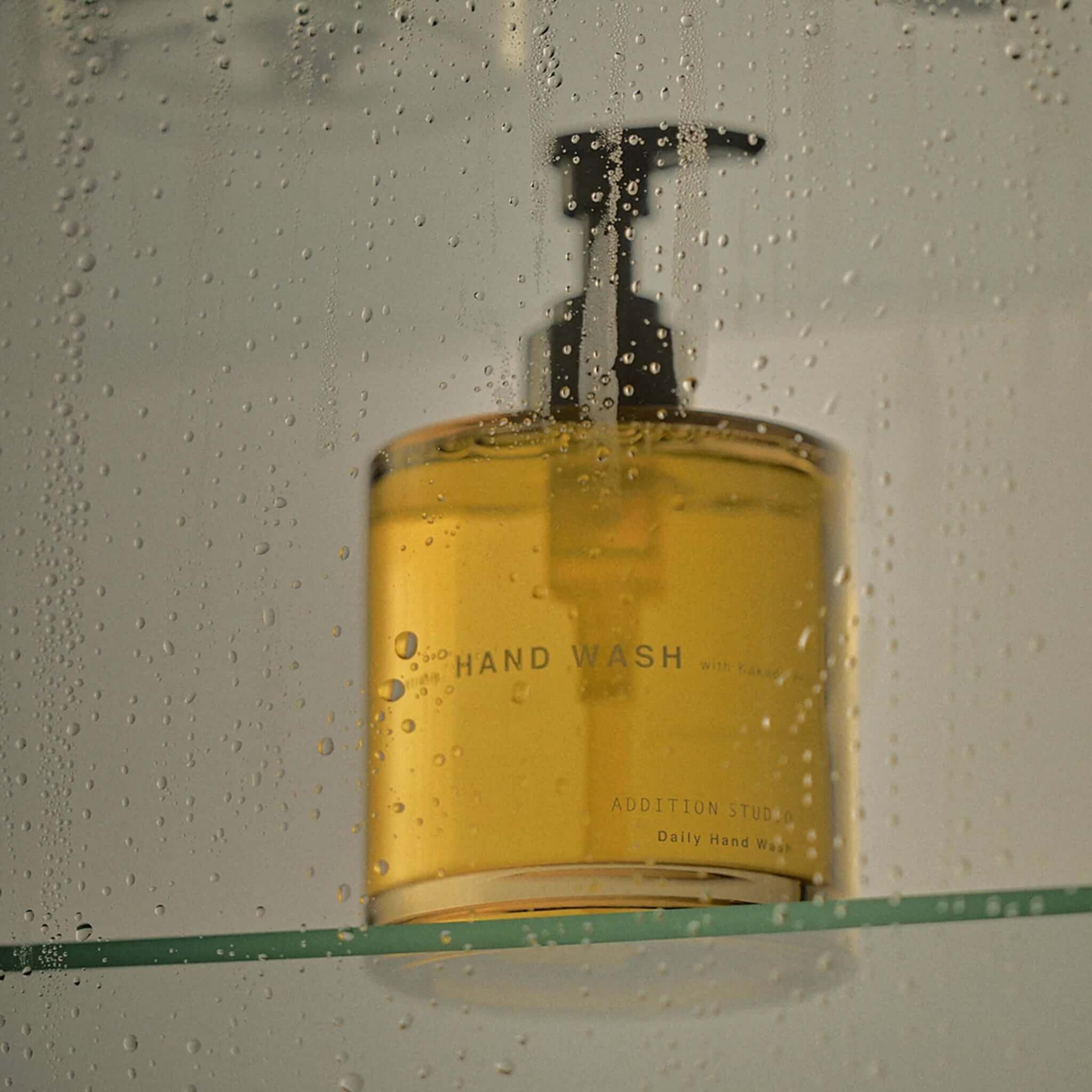 Addition Studio Hand Wash in a glass bottle, with a matte black pump, surrounded by droplets.