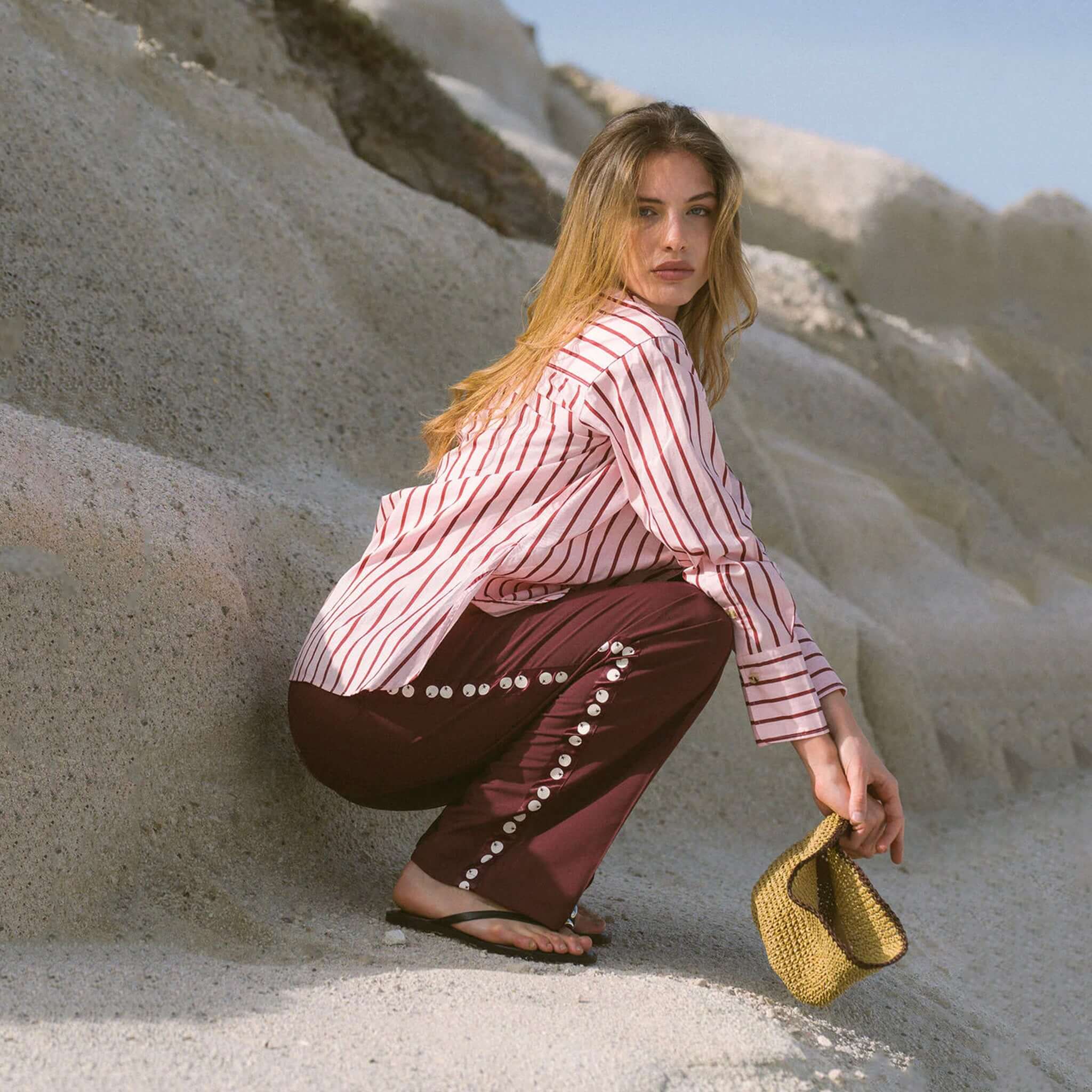 Model wearing Araminta James Pomodoro Twill Pants - Burgundy posed on a rocky beach, showcasing stylish side trims.