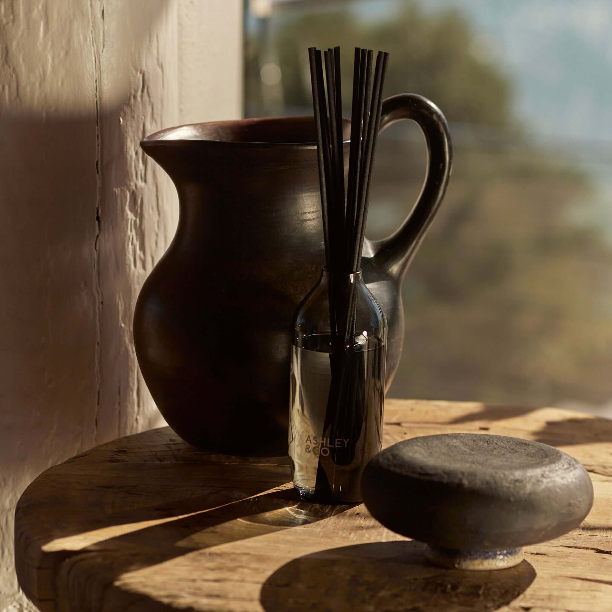 Alt Tag: Ashley & Co Home Perfume Diffusers reed diffuser bottle with black wicks on wooden table beside ceramic vase in warm light