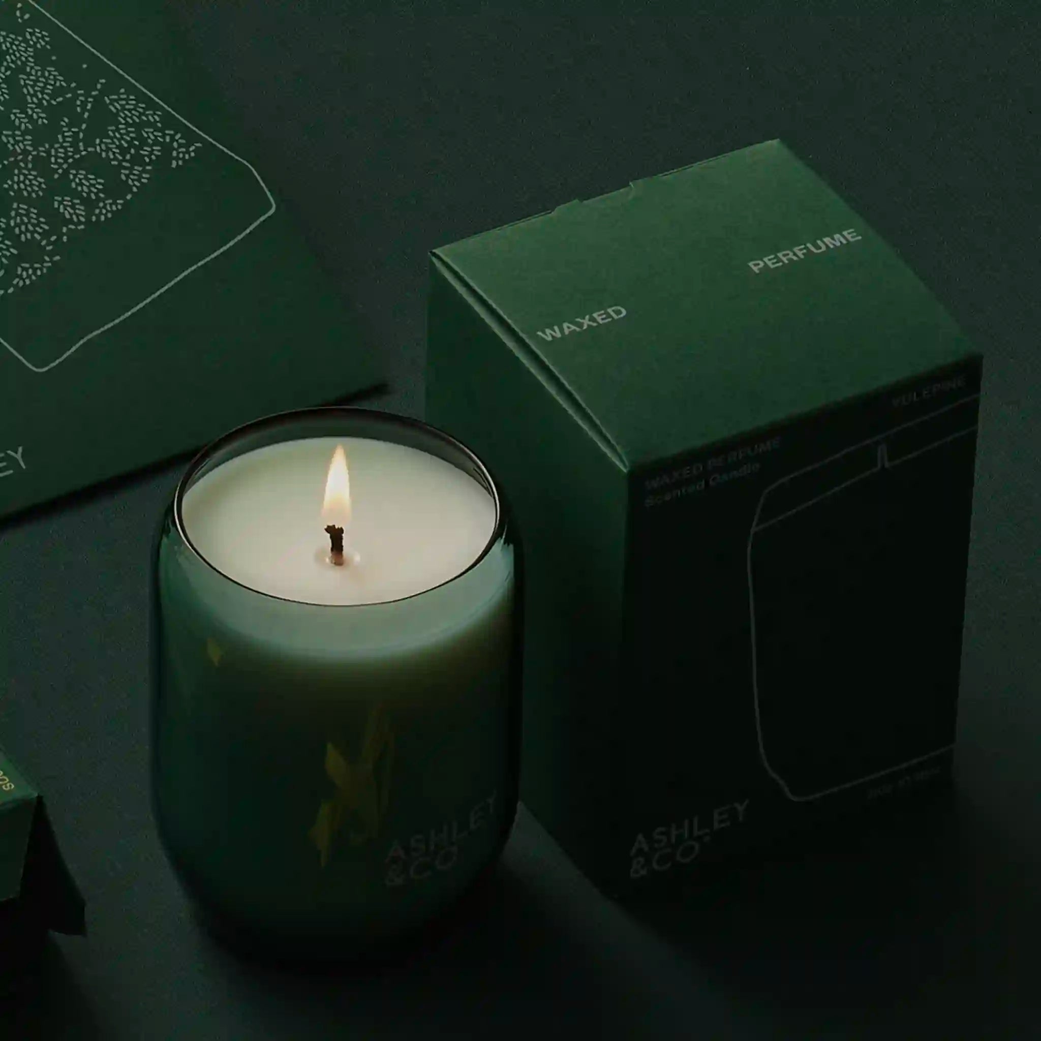 Green candle with a lit wick next to a green box labeled 'Perfume' on a dark background.