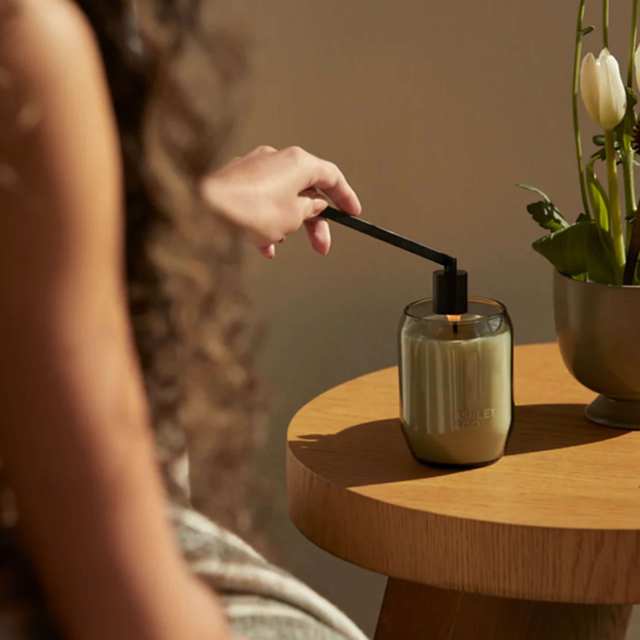 Ashley & Co Snuffy Wick Snuffer extinguishing candle flame over waxed perfume jar, matte black metal 19cm snuffer on wooden table