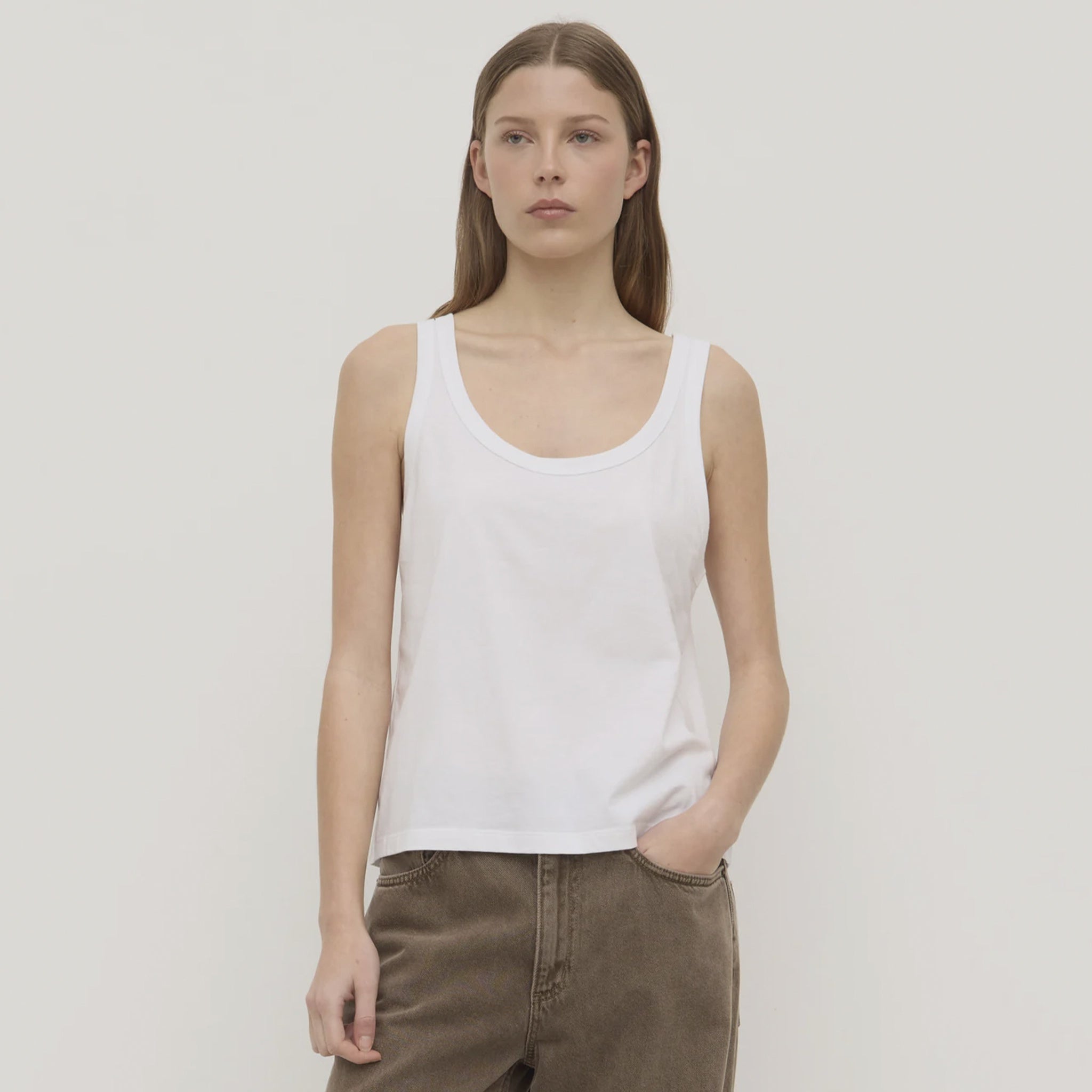 Woman wearing a white tank top and brown pants against a plain background