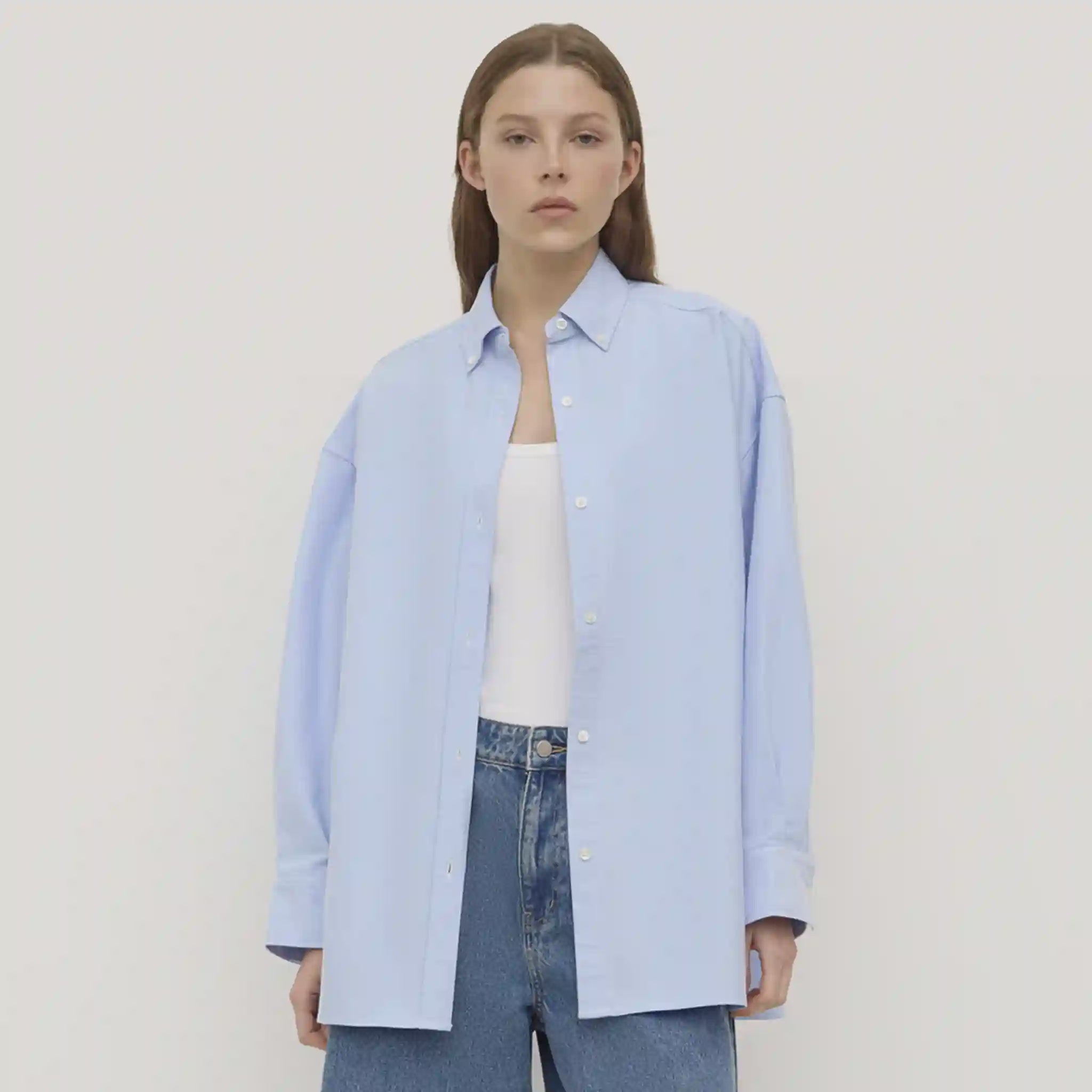 Person wearing a light blue shirt over a white top with blue jeans on a plain background