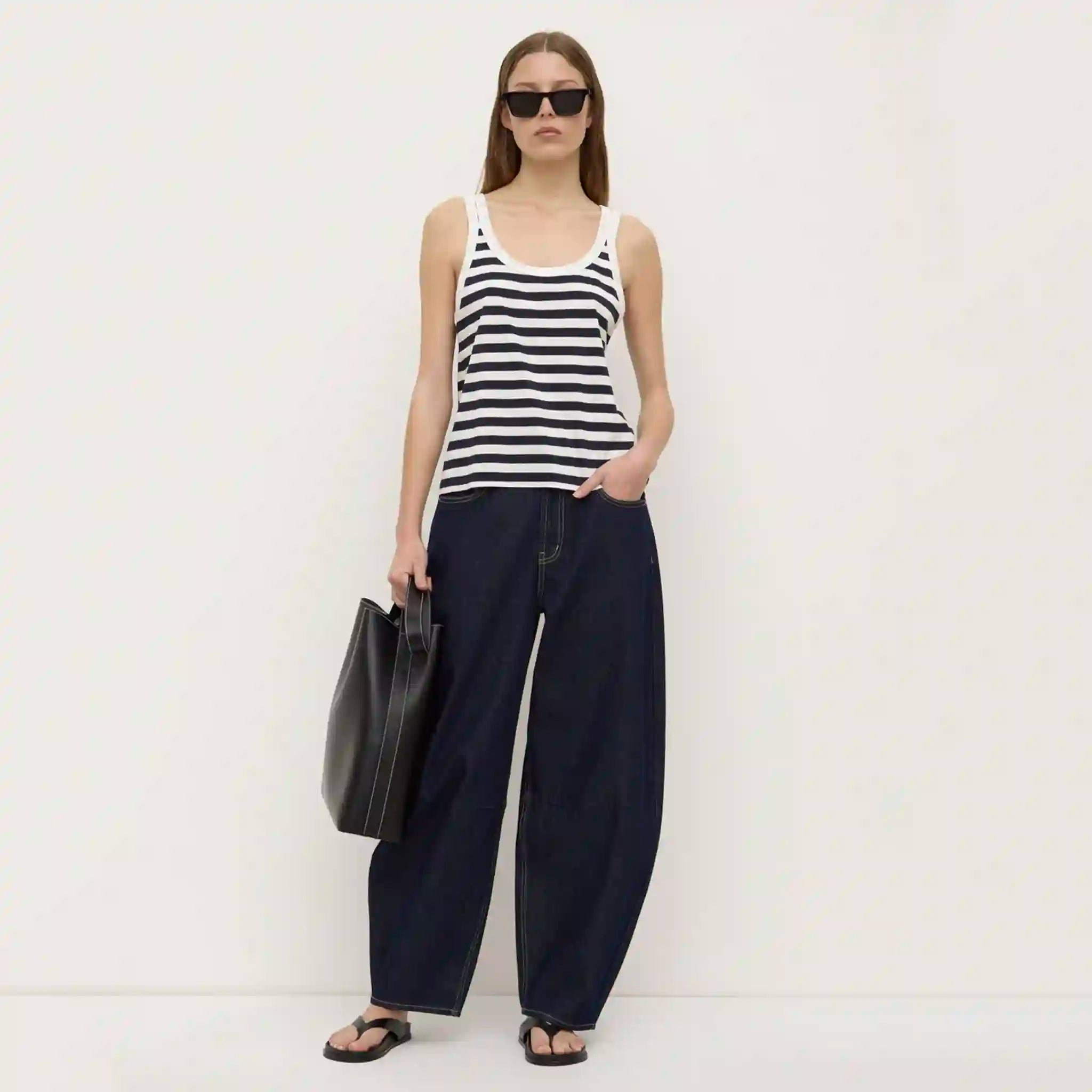 Woman wearing a black and white striped tank top and navy pants, holding a black bag on a white background