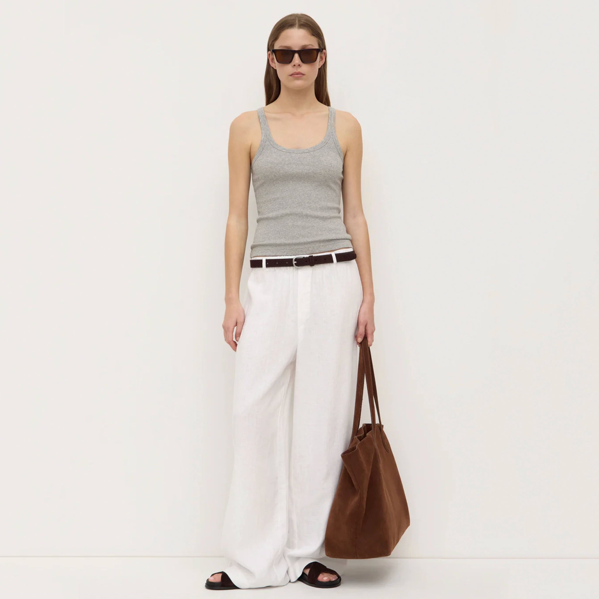 Woman wearing a gray tank top and white pants, holding a brown bag on a white background