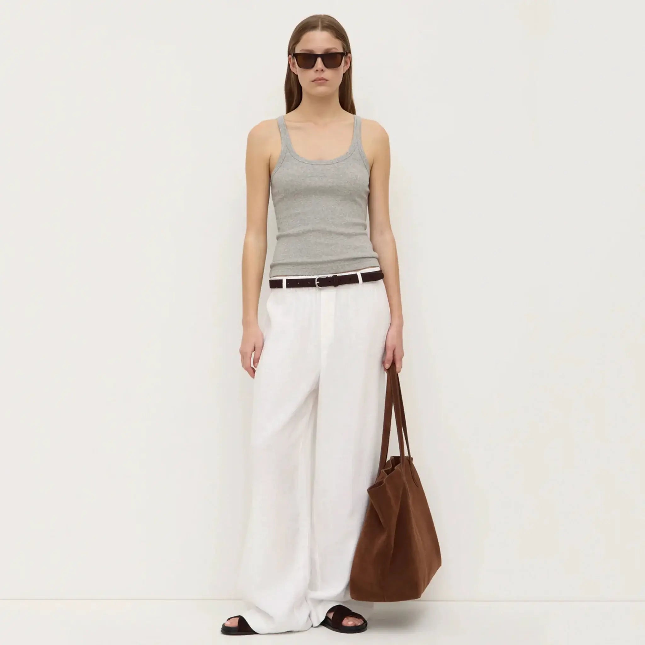 Woman wearing a gray tank top and white pants, holding a brown bag on a white background