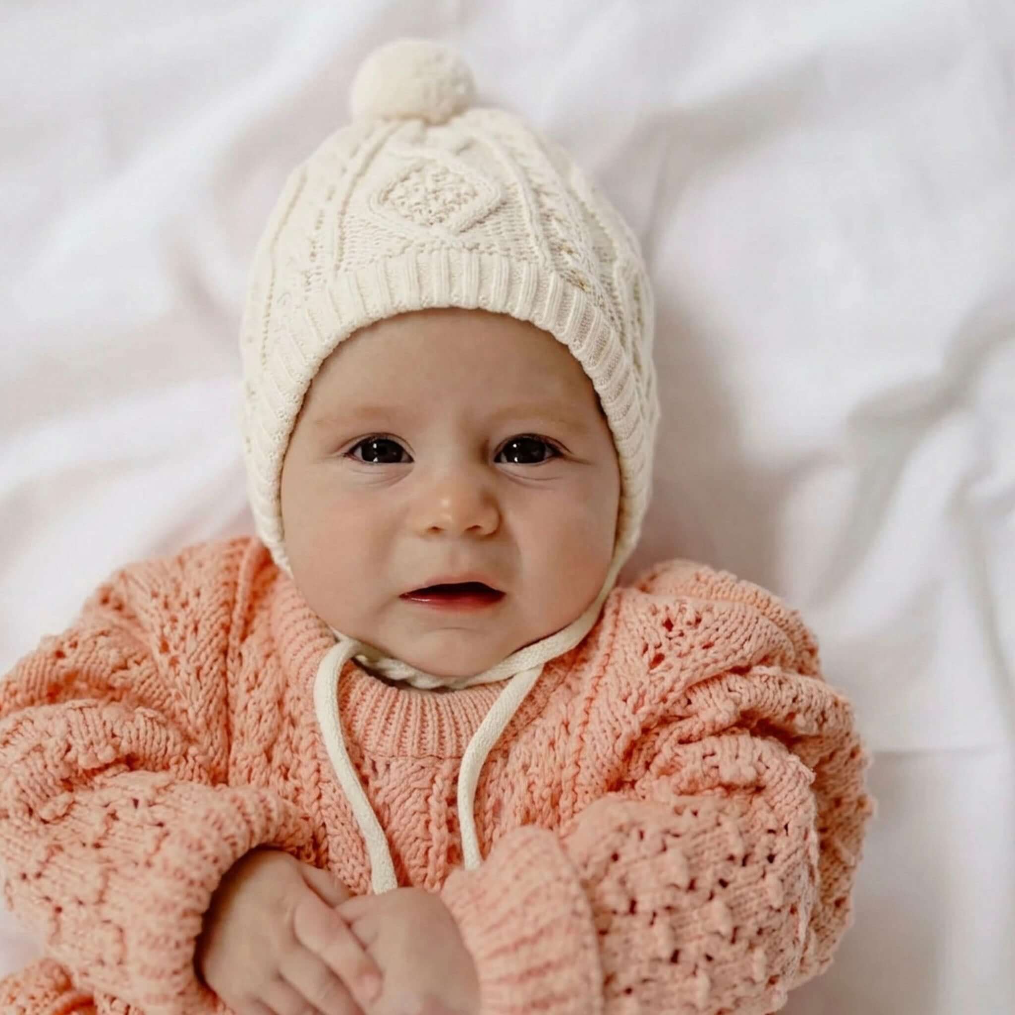 Baby Cotton Luna Beanie - Ivory worn by an infant with a cozy sweater, showcasing its soft cable knit design.