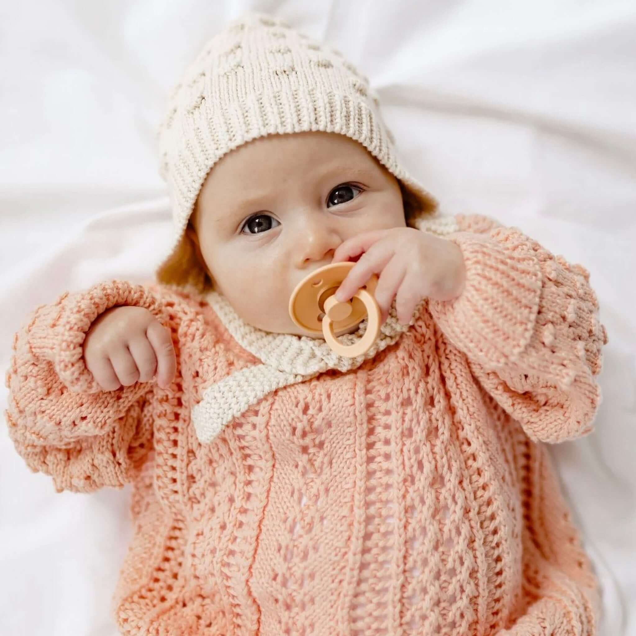 Baby Cotton Marshmallow Bonnet - Ivory worn by an infant, pacifier in mouth, dressed in a cozy knit outfit.