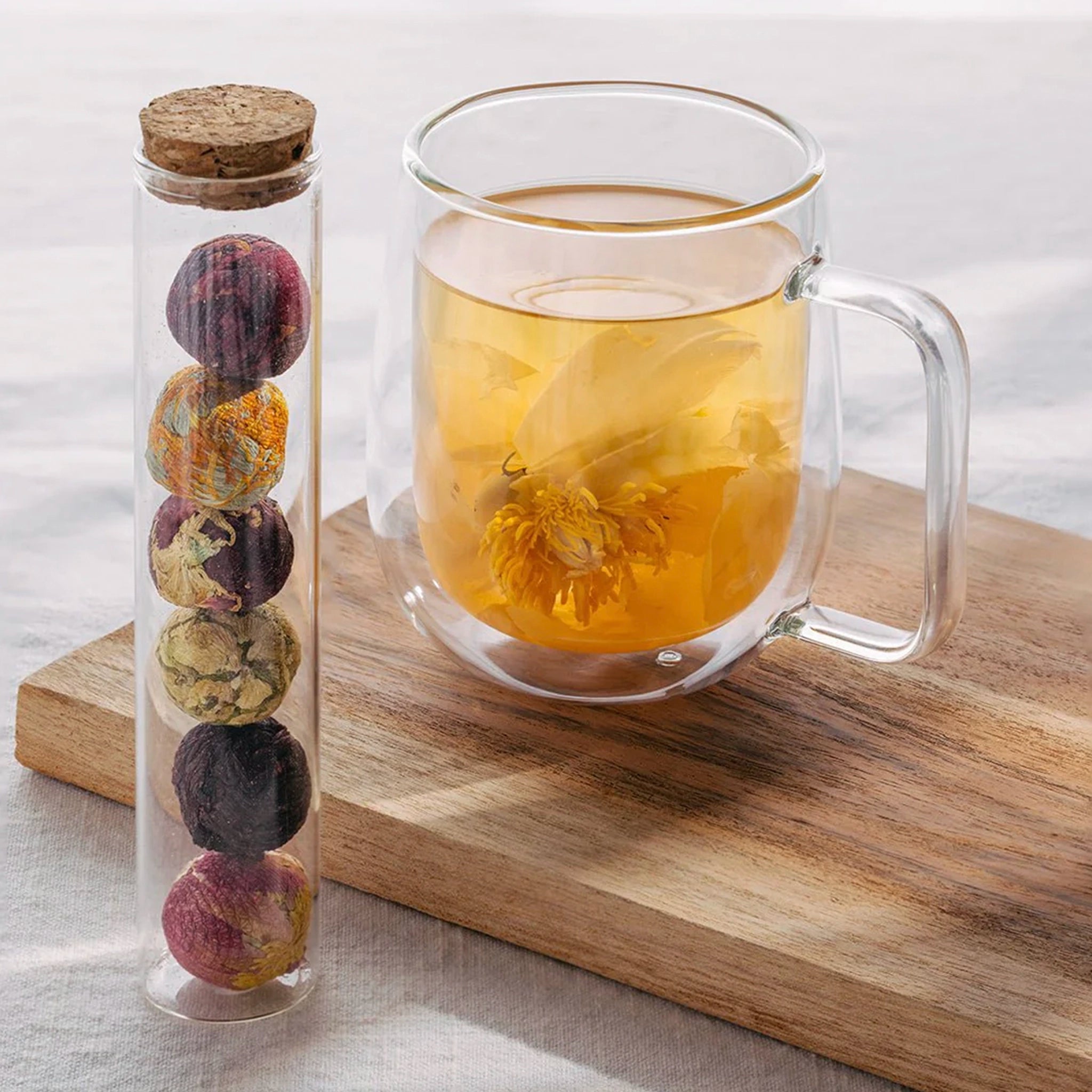 Glass container with dried tea balls next to a glass mug of hot tea on a wooden board.