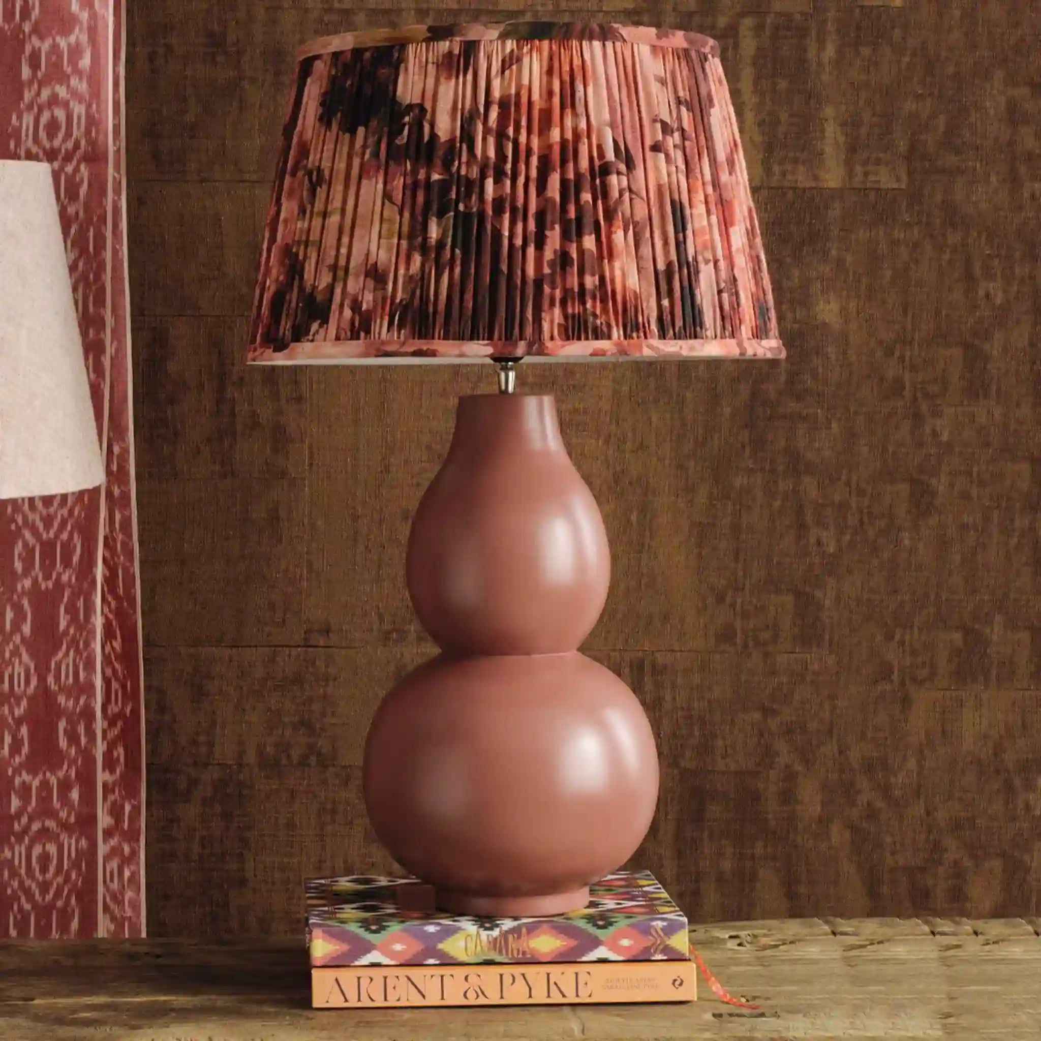 Pink lamp with a patterned shade on a wooden surface against a brown textured wall.