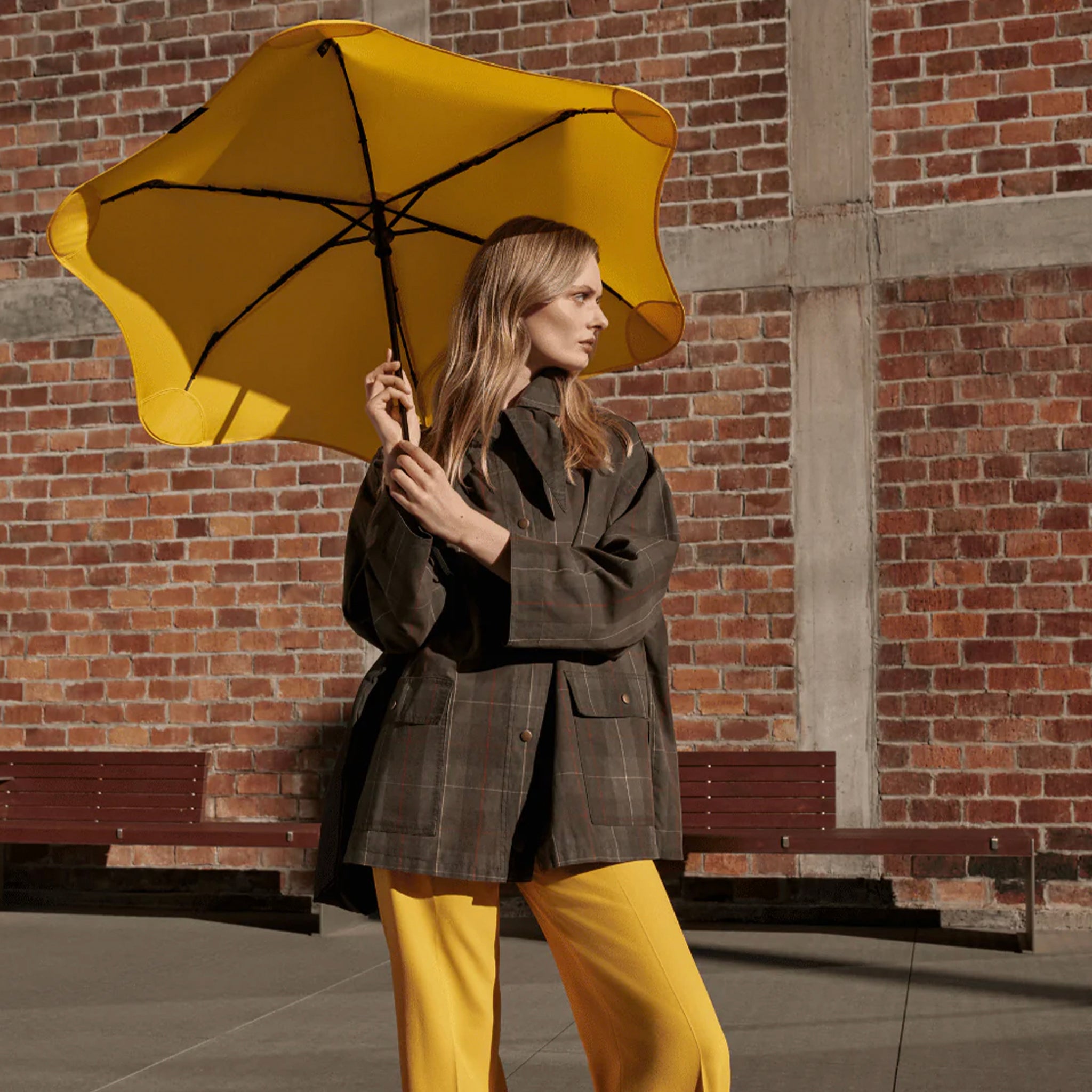 Woman holding Blunt Neo Umbrella - Citrus Yellow against a brick wall, showcasing its stylish design.