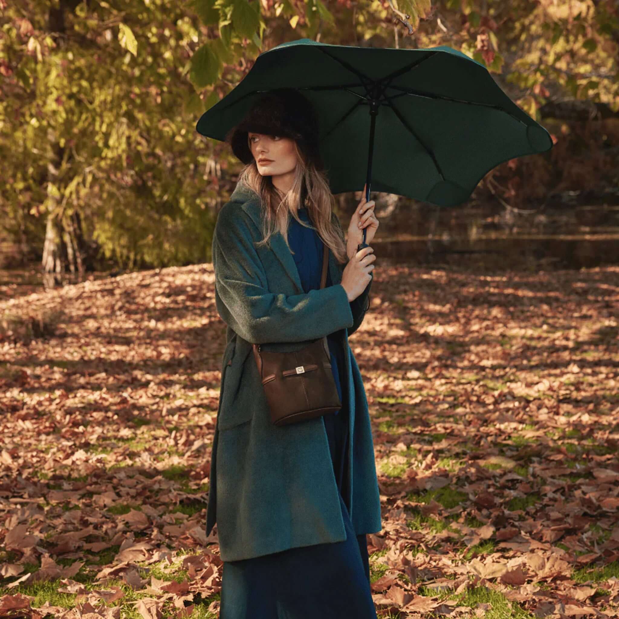 Woman holding Blunt Neo Umbrella - Forest Green in a forest setting with autumn leaves.