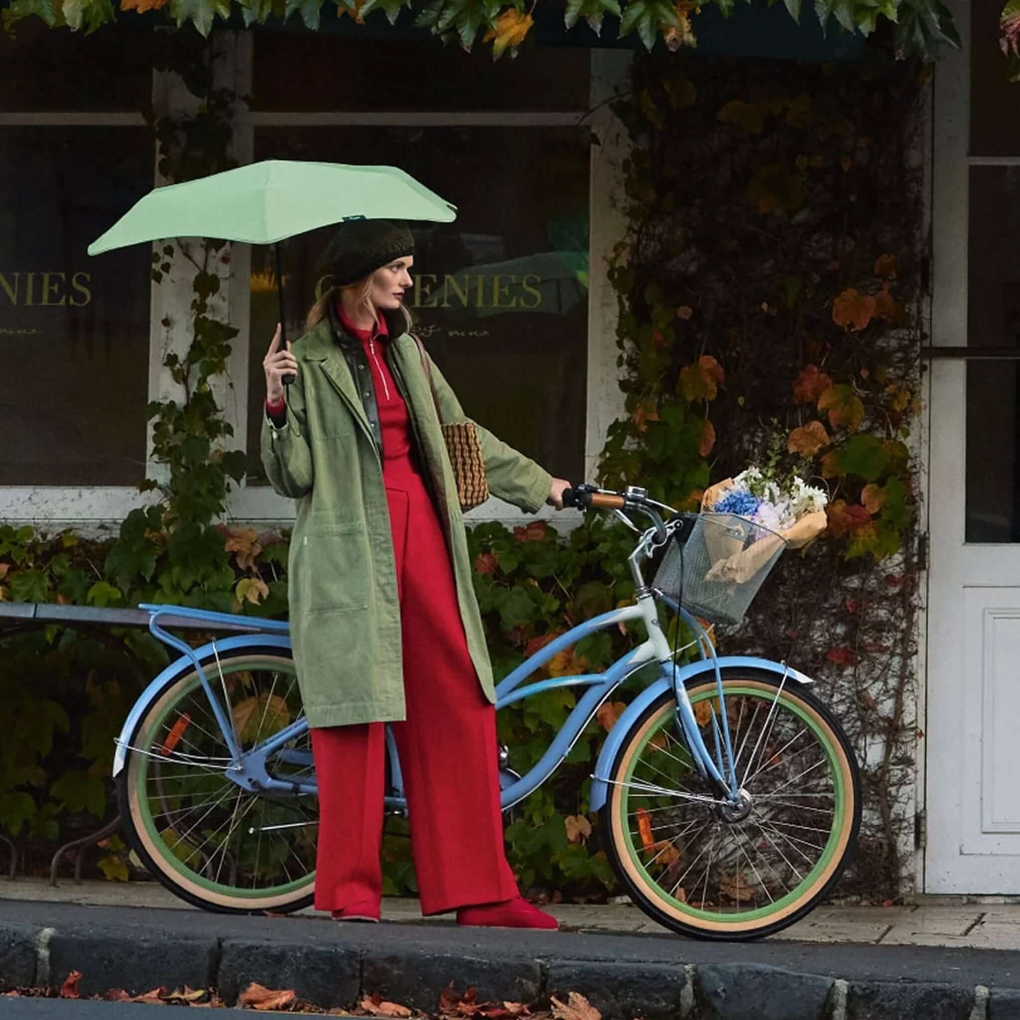 Woman in green coat holding Blunt Neo Umbrella - Sage Green while standing next to a bicycle with flowers.