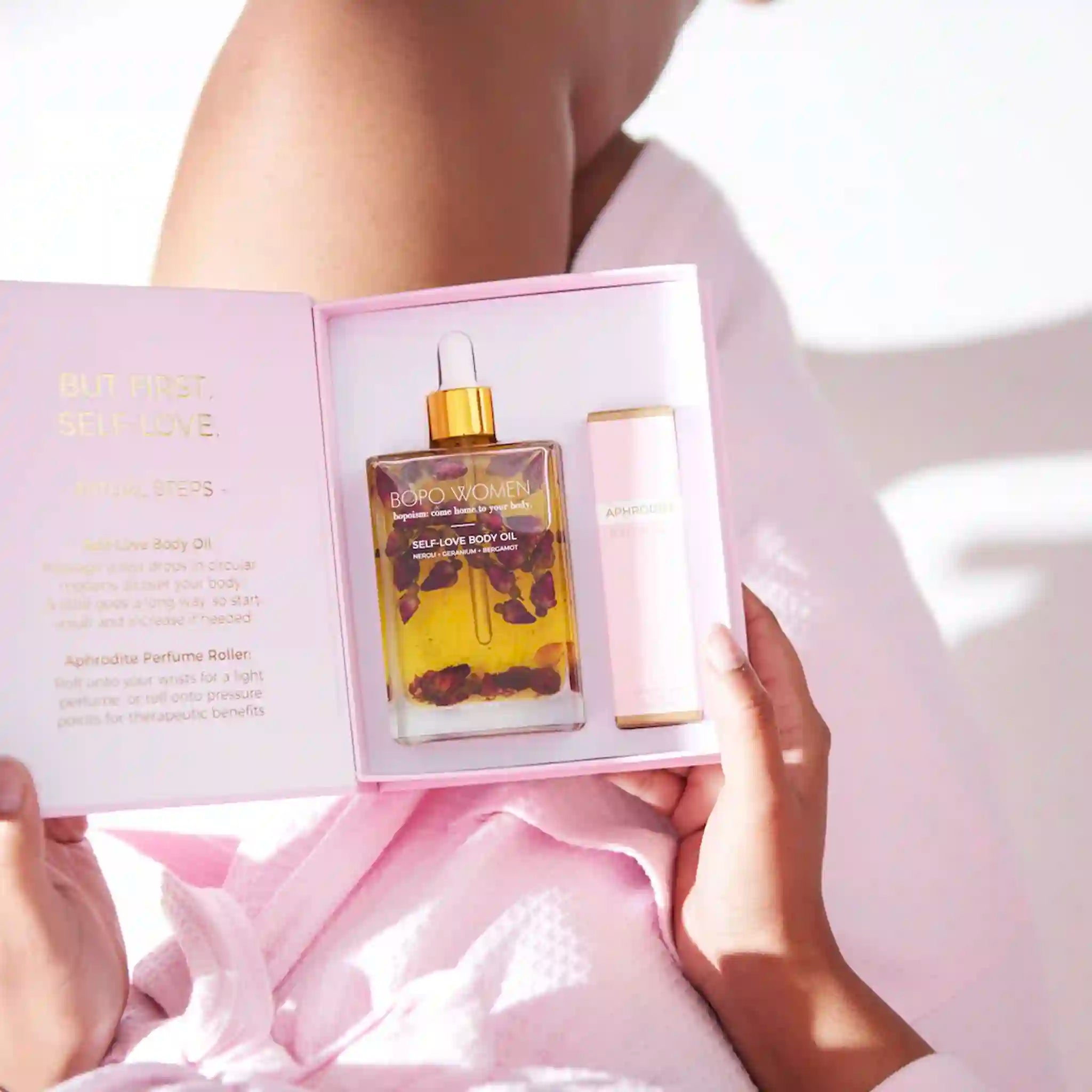 Person holding a box with a bottle of body oil and a pink container inside, against a light background.
