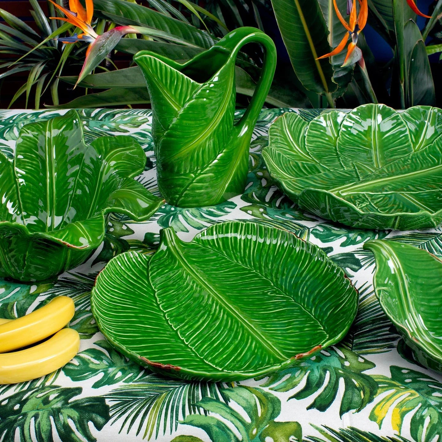 Green leaf-patterned ceramic items on a matching tablecloth with tropical plants in the background.