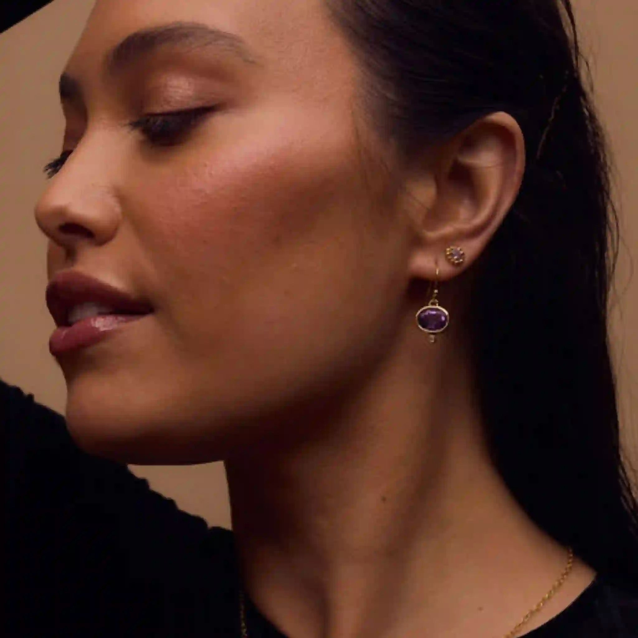 Close-up of a woman wearing a gold earring with a purple gemstone on a neutral background