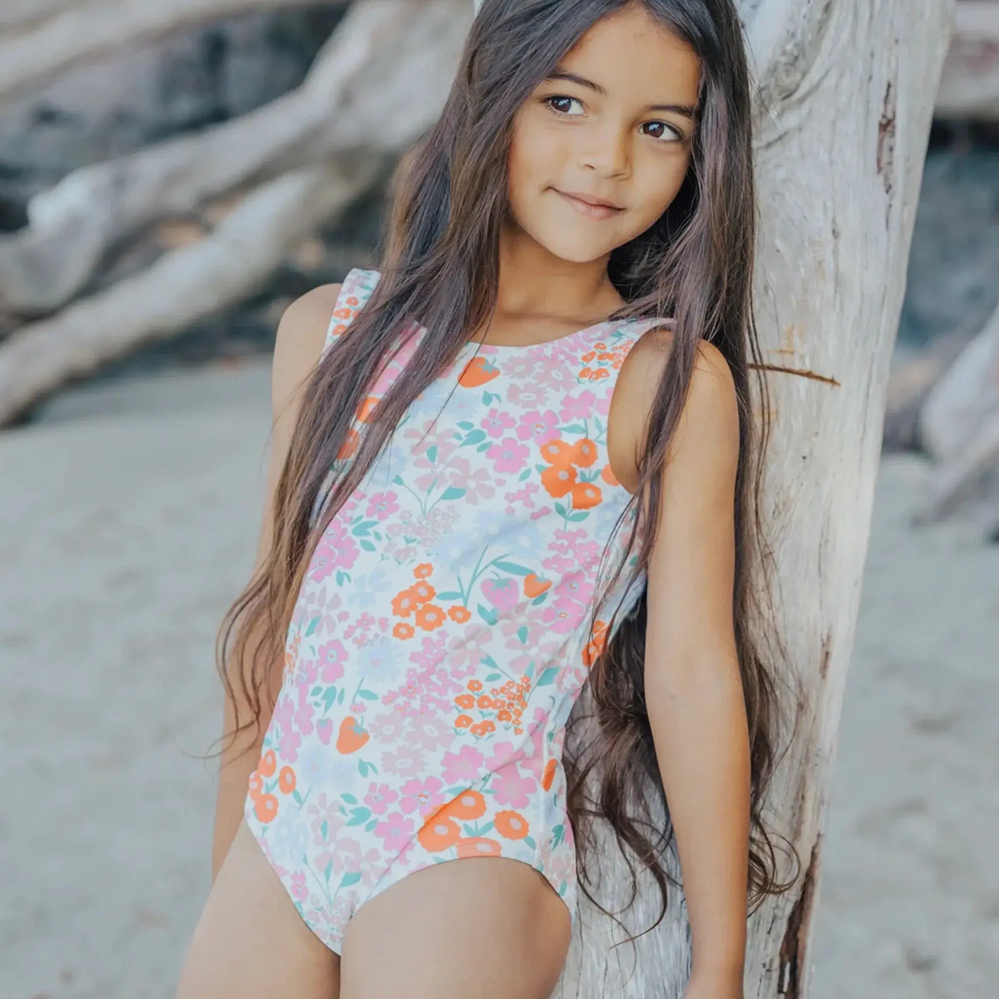 Young girl in a floral swimsuit standing next to a tree on a beach.