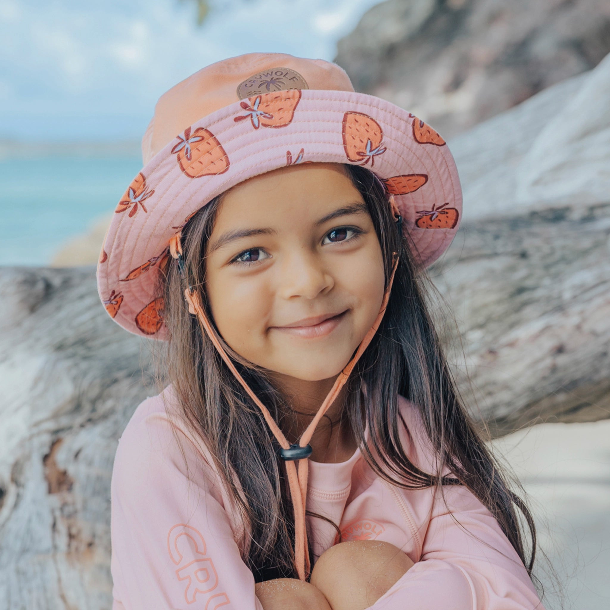 Young girl wearing a pink bucket hat with strawberry pattern sitting on a log by a body of water.