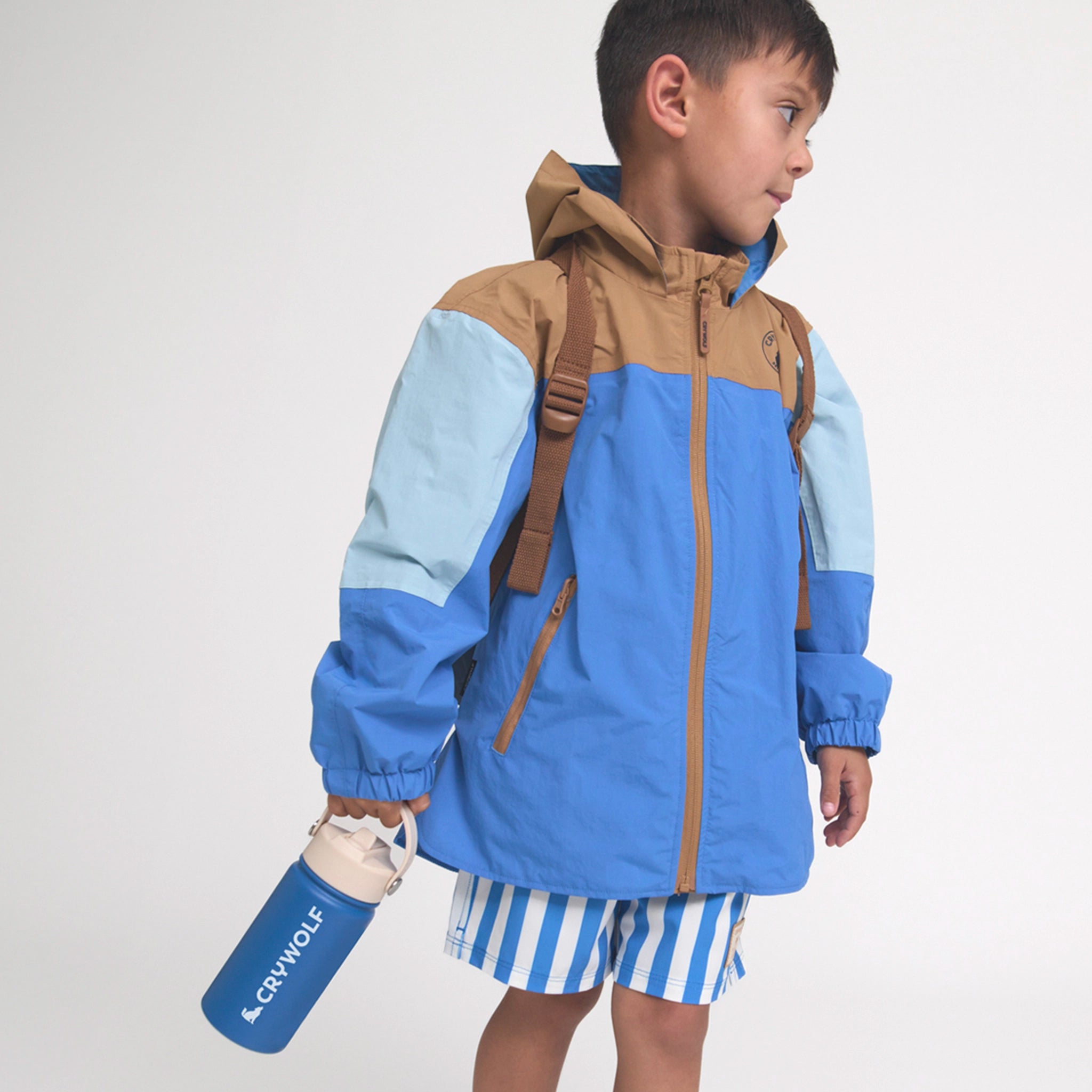 Child wearing a blue and brown jacket with a backpack, holding a water bottle on a white background