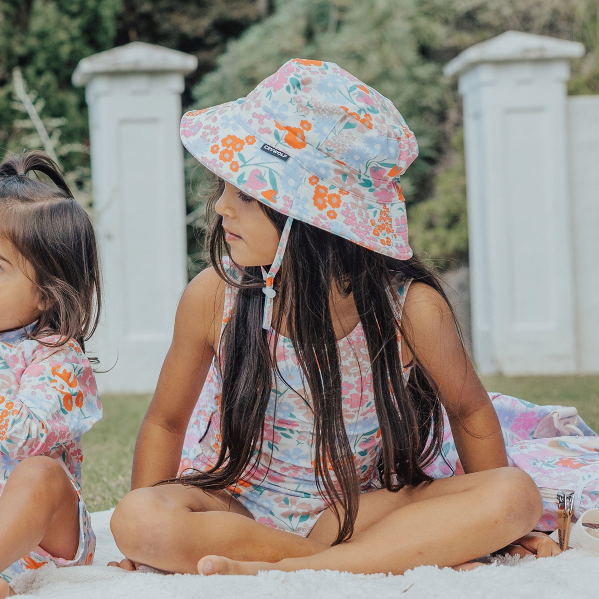Two young girls sitting on a blanket outdoors, wearing floral outfits and hats.