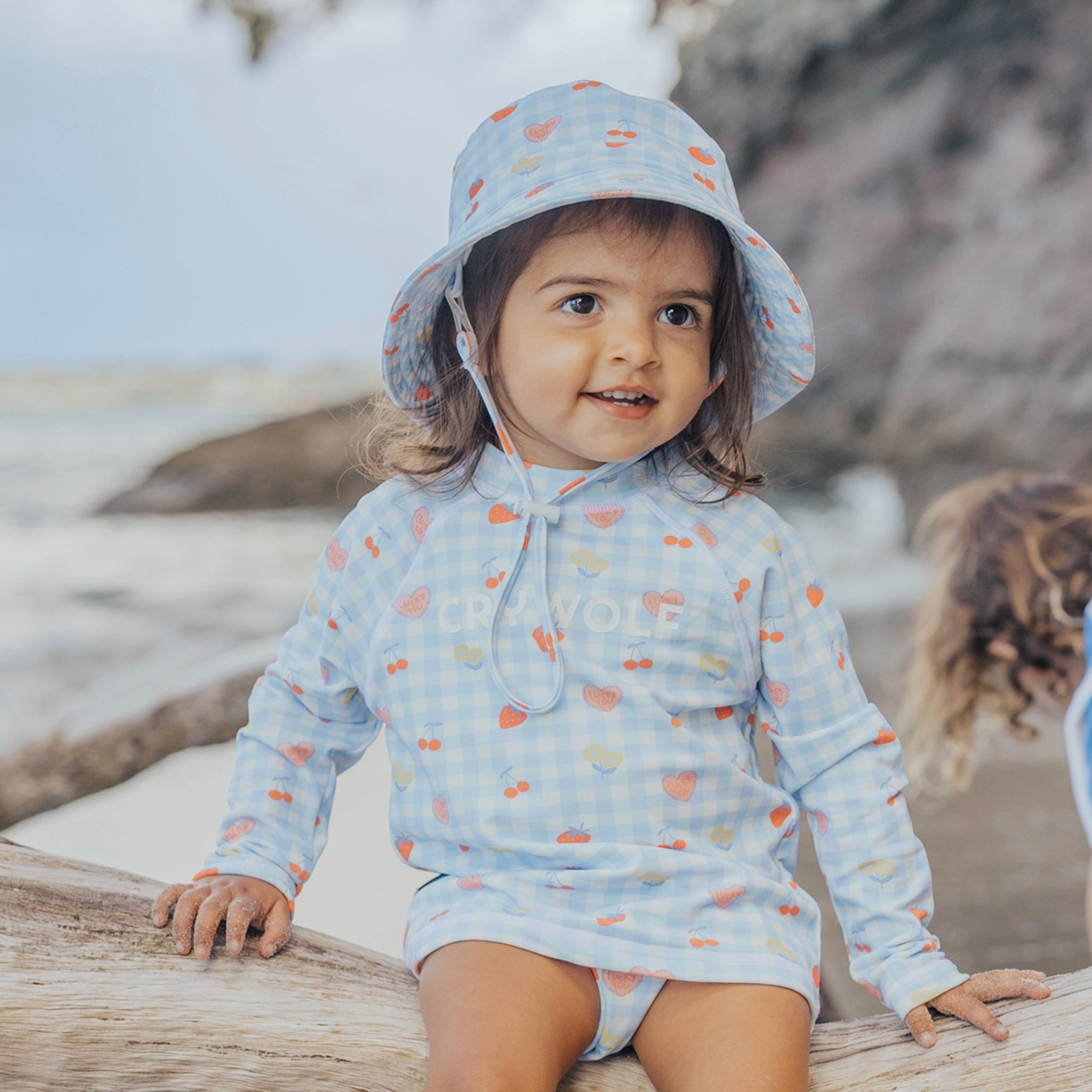 Child wearing a light blue hoodie and hat with colorful patterns, sitting on a log at the beach.