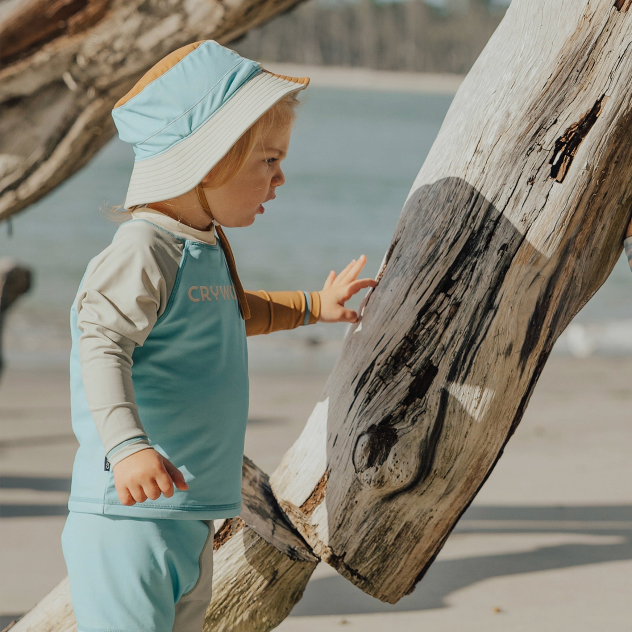 Child in light blue outfit and sun hat touching a large wooden log on a beach.