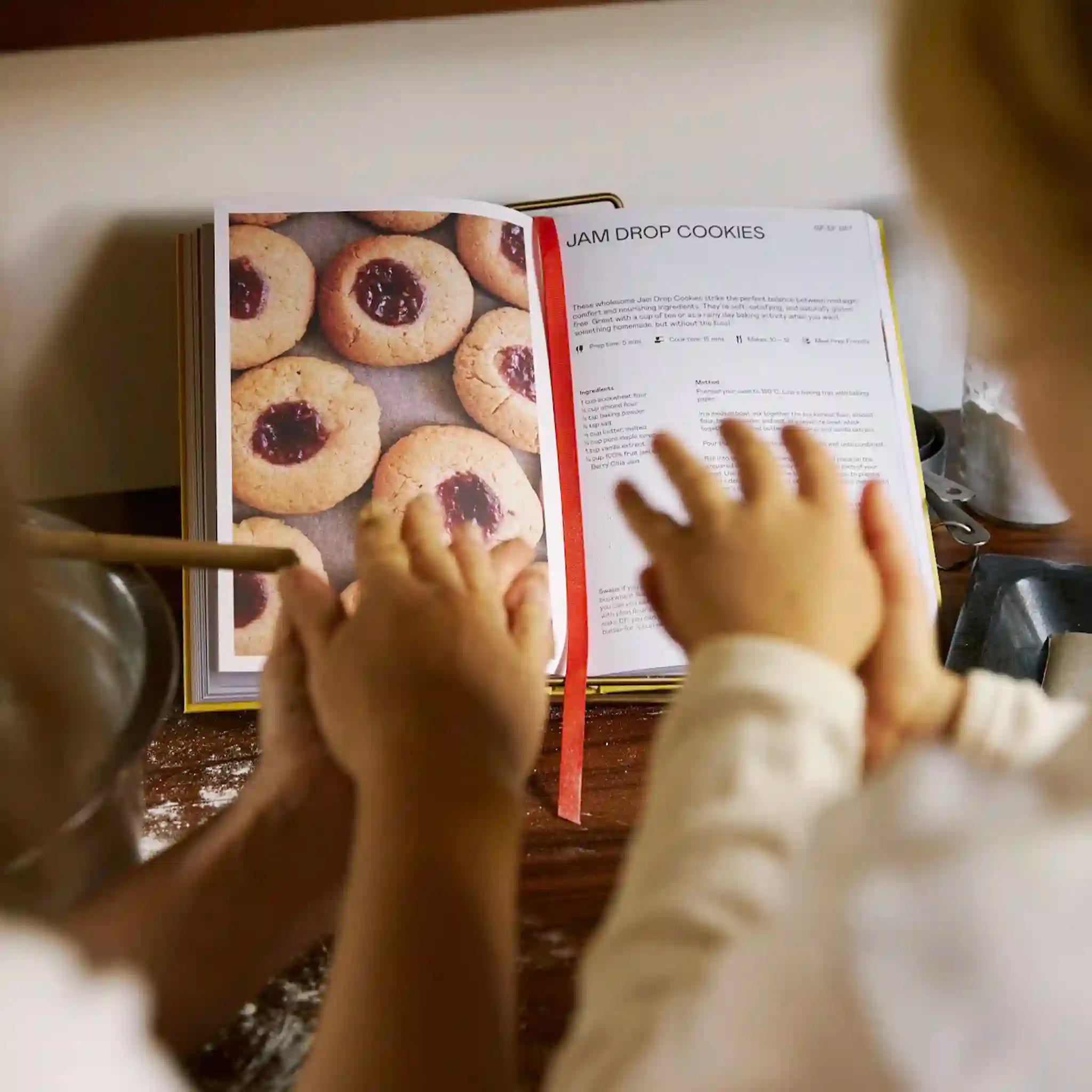 Two children's hands reaching towards a cookbook open to a page about jam drop cookies.