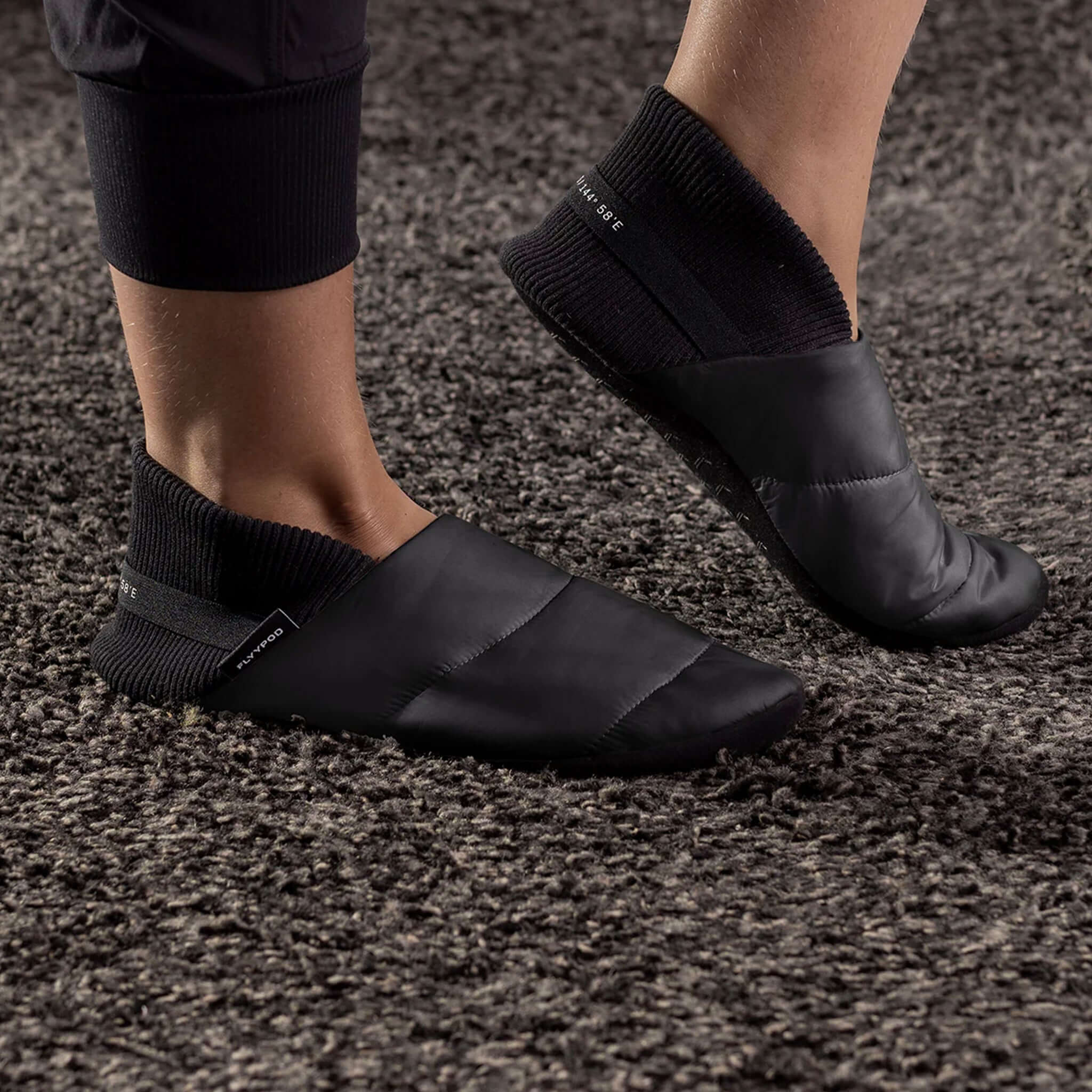 Alt Tag: Flyypod Skyywalkers Travel Slippers - Tarmac worn on feet, black puffer travel slippers with ribbed ankle cuff on carpet