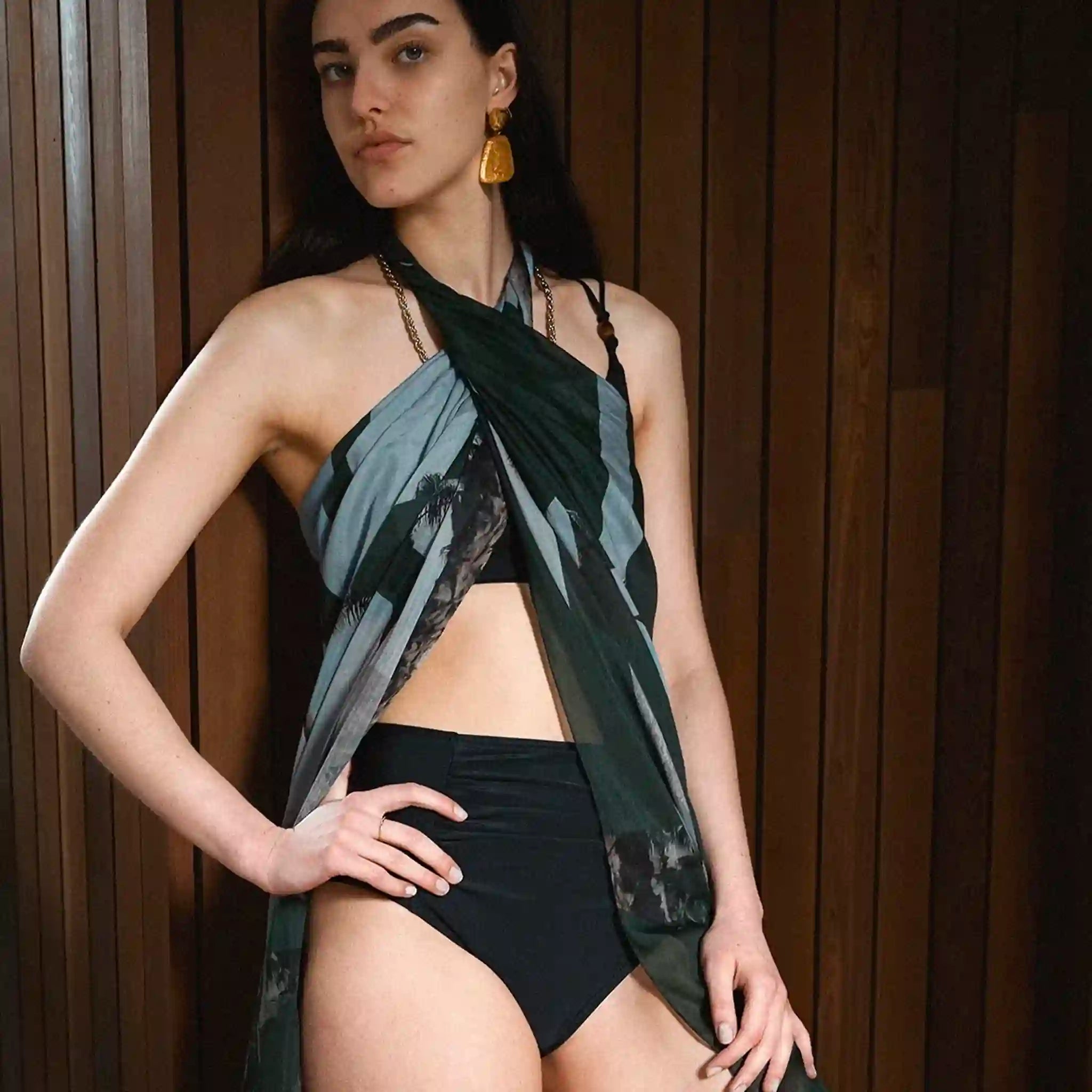 Woman wearing a black halter top and high-waisted black bikini bottom with a green and blue patterned cover-up against a wooden background.