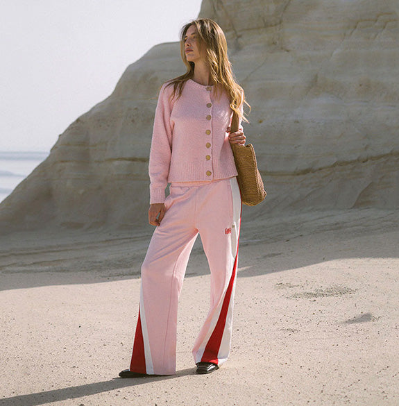 A woman stands on a sandy beach in front of a large pale rock formation, wearing a light pink knit cardigan with gold buttons and matching wide-leg pink pants with red side panels. She is holding a woven tan bag.