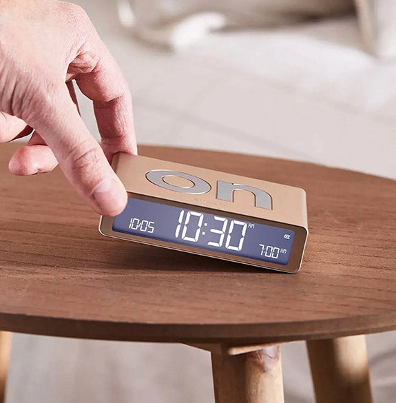 Digital clock on a wooden surface with a hand pressing a button.