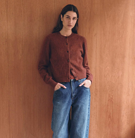 Woman wearing a brown sweater and blue jeans against a wooden wall