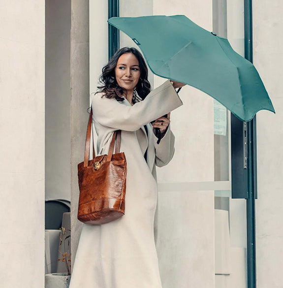 Woman holding a teal umbrella and brown leather handbag in an urban setting