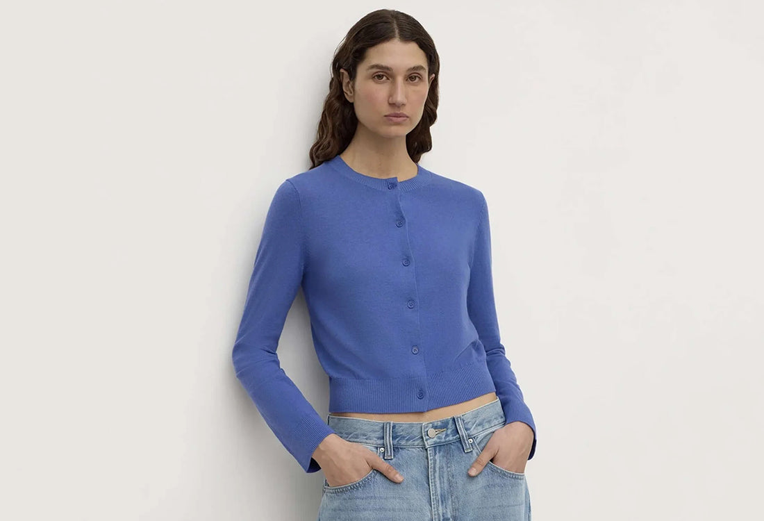 A woman modeling a blue long-sleeve cropped button-front cardigan with light-wash blue jeans, standing against a plain off-white wall.