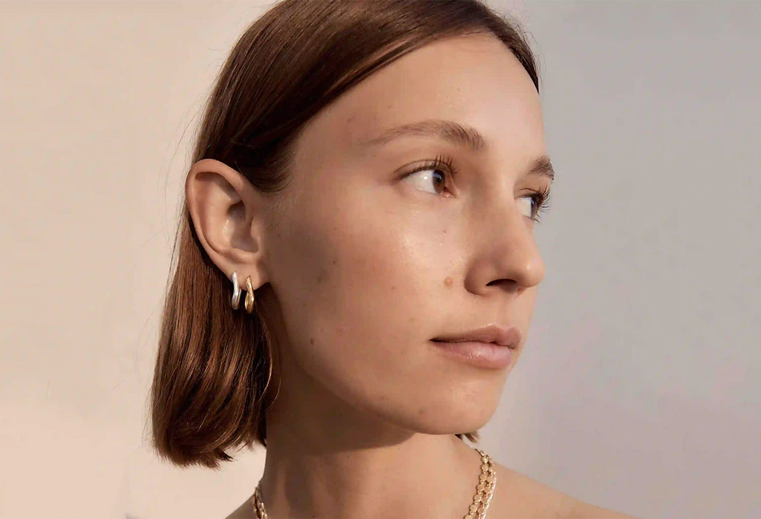 Close-up of a woman wearing gold earrings and a necklace against a neutral background