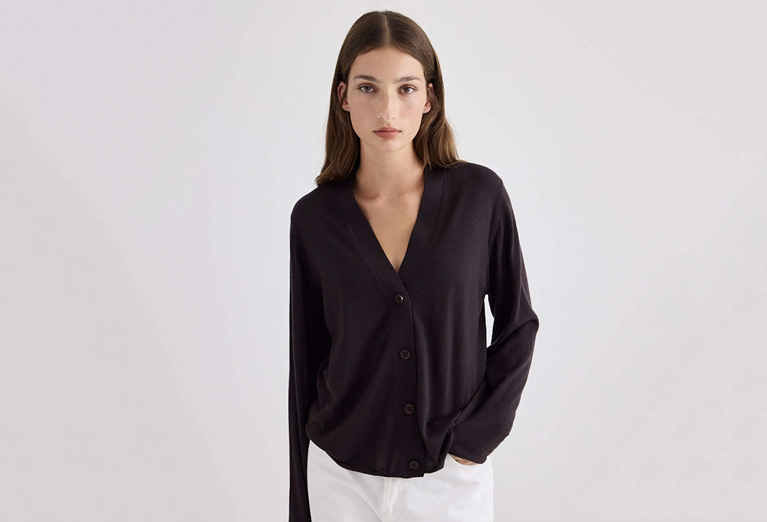 A woman modeling a black long-sleeve V-neck button-up cardigan top with white pants, photographed against a clean light gray studio background.