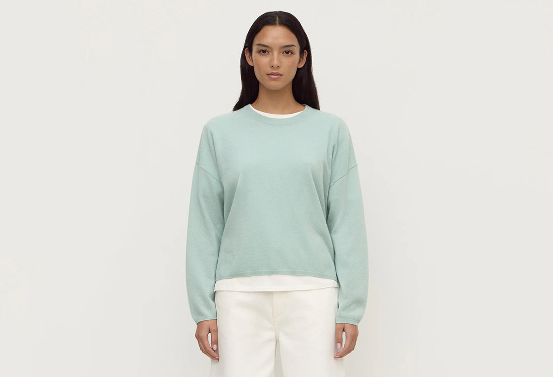 Woman wearing a light green sweater and white pants on a plain background