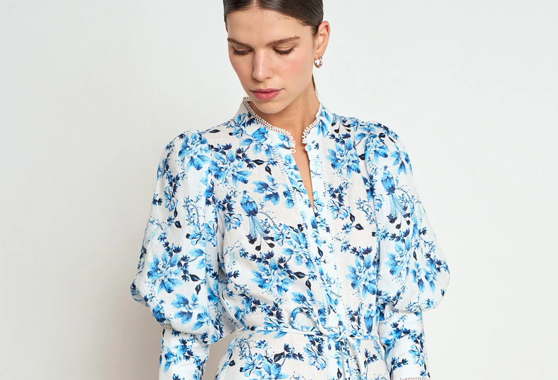 A woman wearing a white long-sleeve blouse with a blue floral print, featuring a high collar and puffed sleeves, photographed against a plain light gray background.