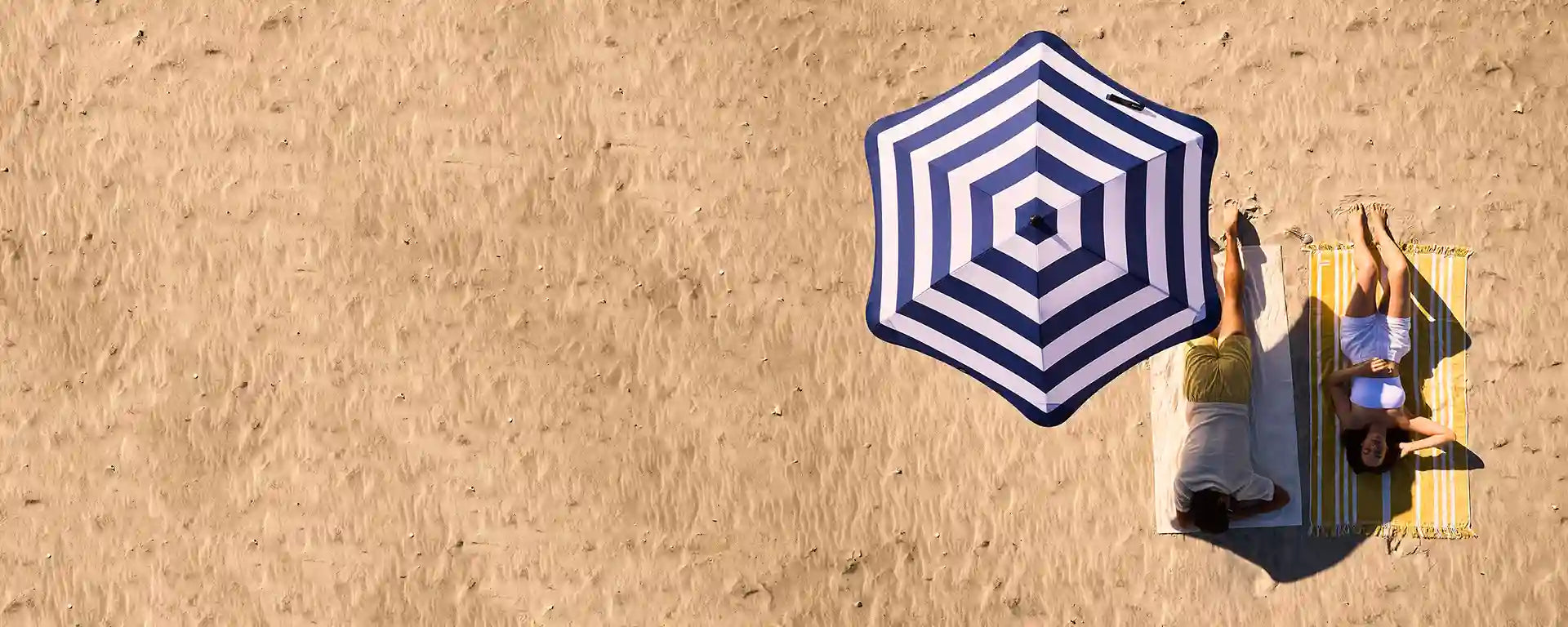 Two people lying on towels under a blue and white striped umbrella on a sandy beach.