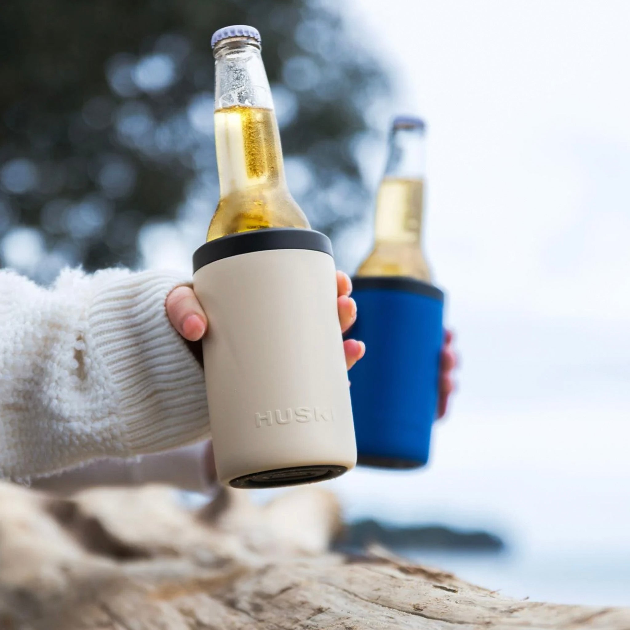 Two bottles of beer in Huski bottle coolers held by hands against a blurred natural background.
