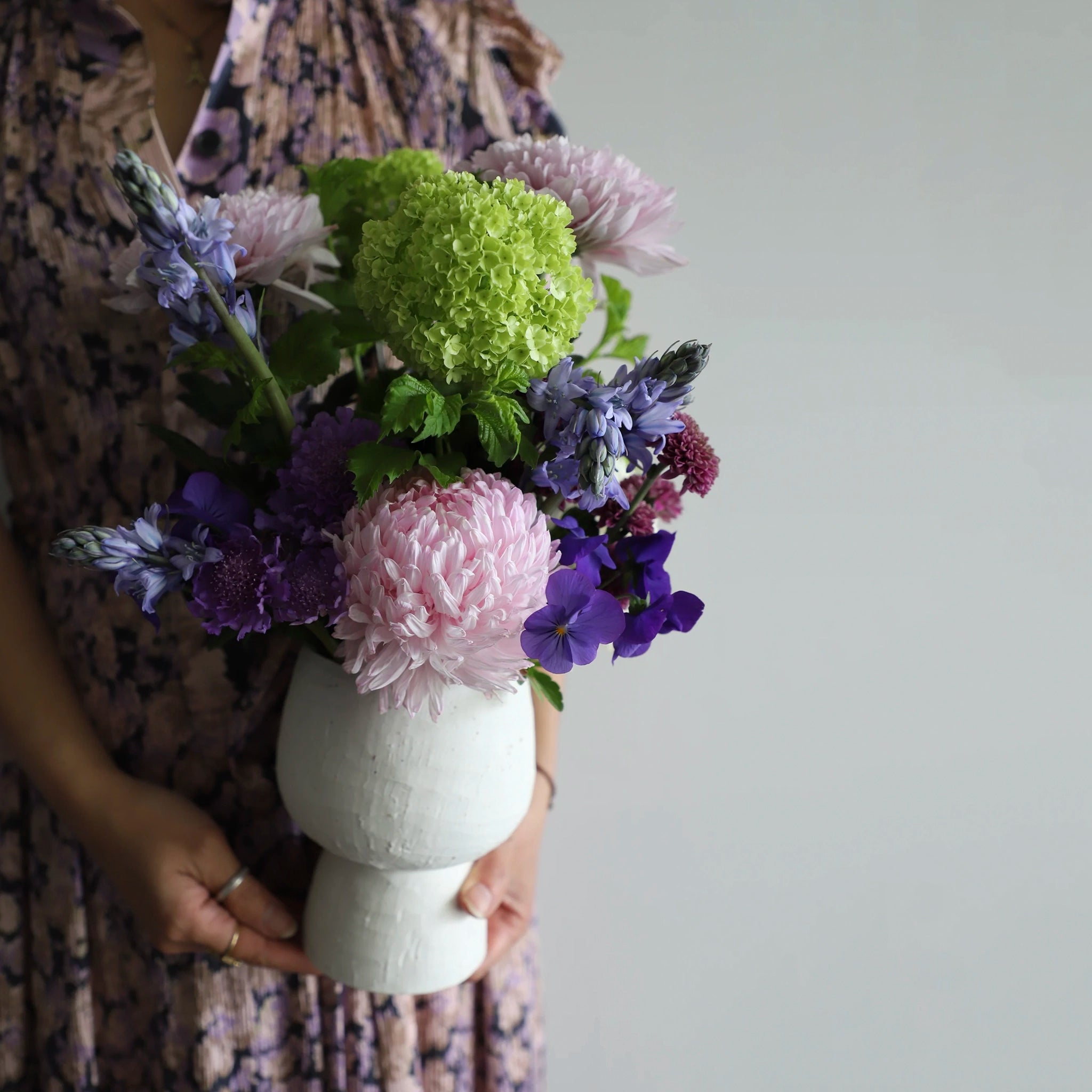 Person holding a bouquet of flowers in a white vase against a plain background