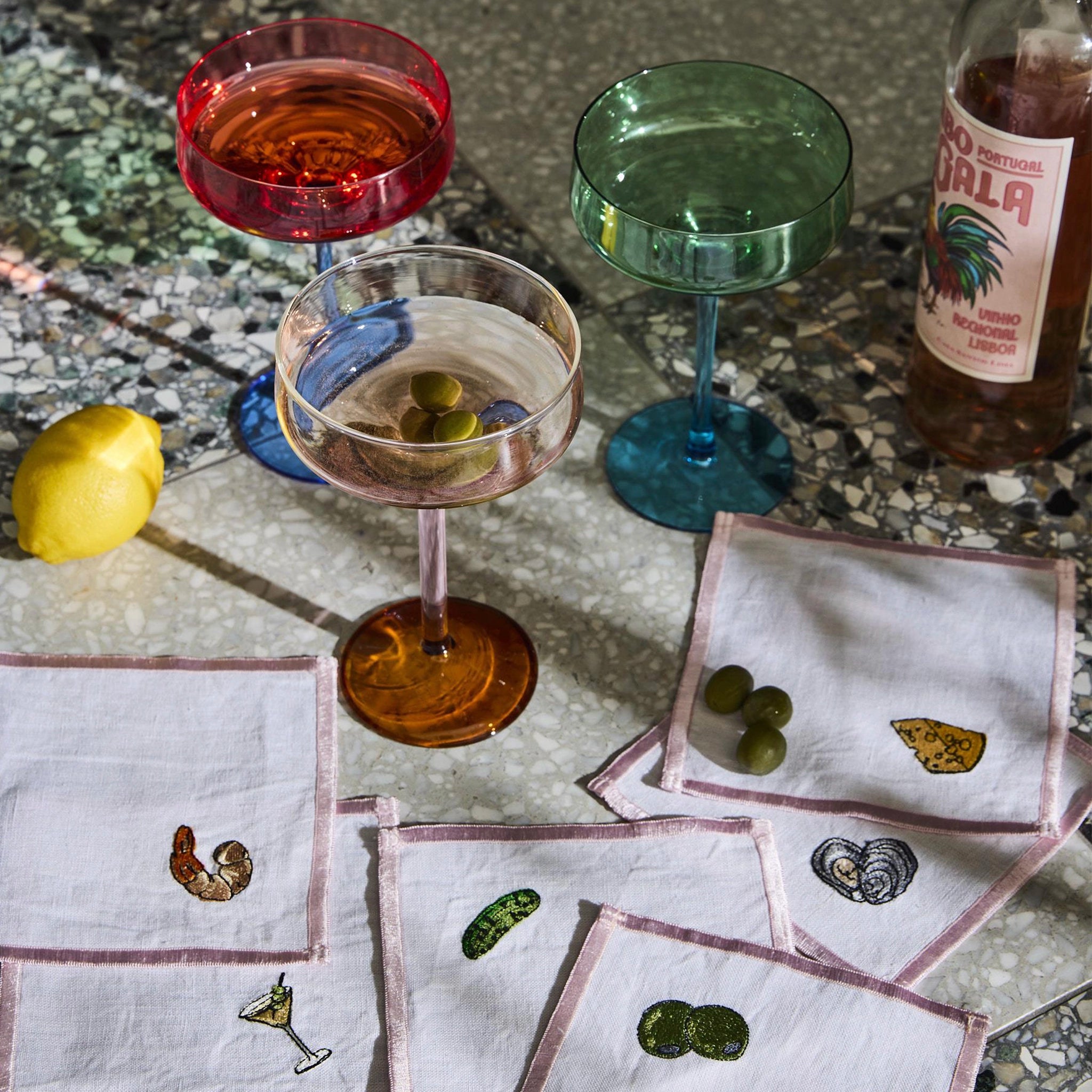 Cocktail glasses with colored liquids on a textured surface with bottles and garnishes.