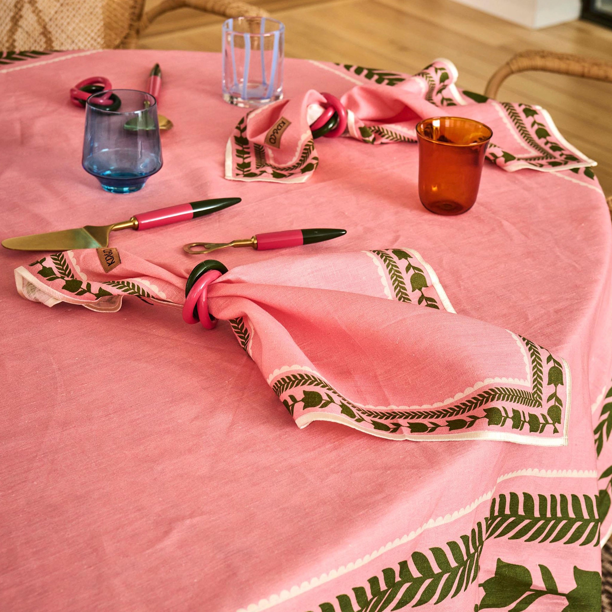 Pink tablecloth with green leaf patterns on a wooden table with glasses and cutlery.