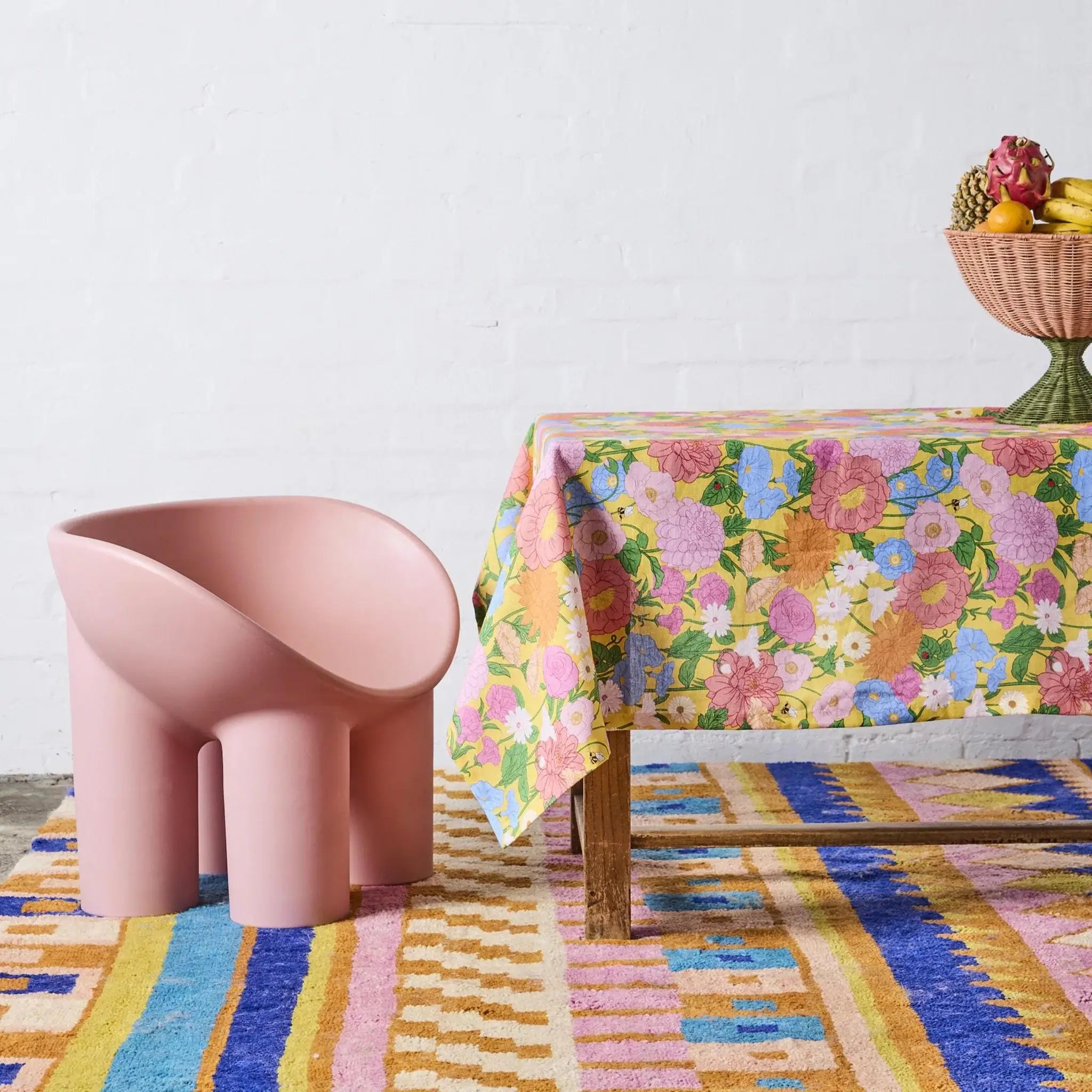 Pink chair next to a table with a floral tablecloth on a colorful rug.