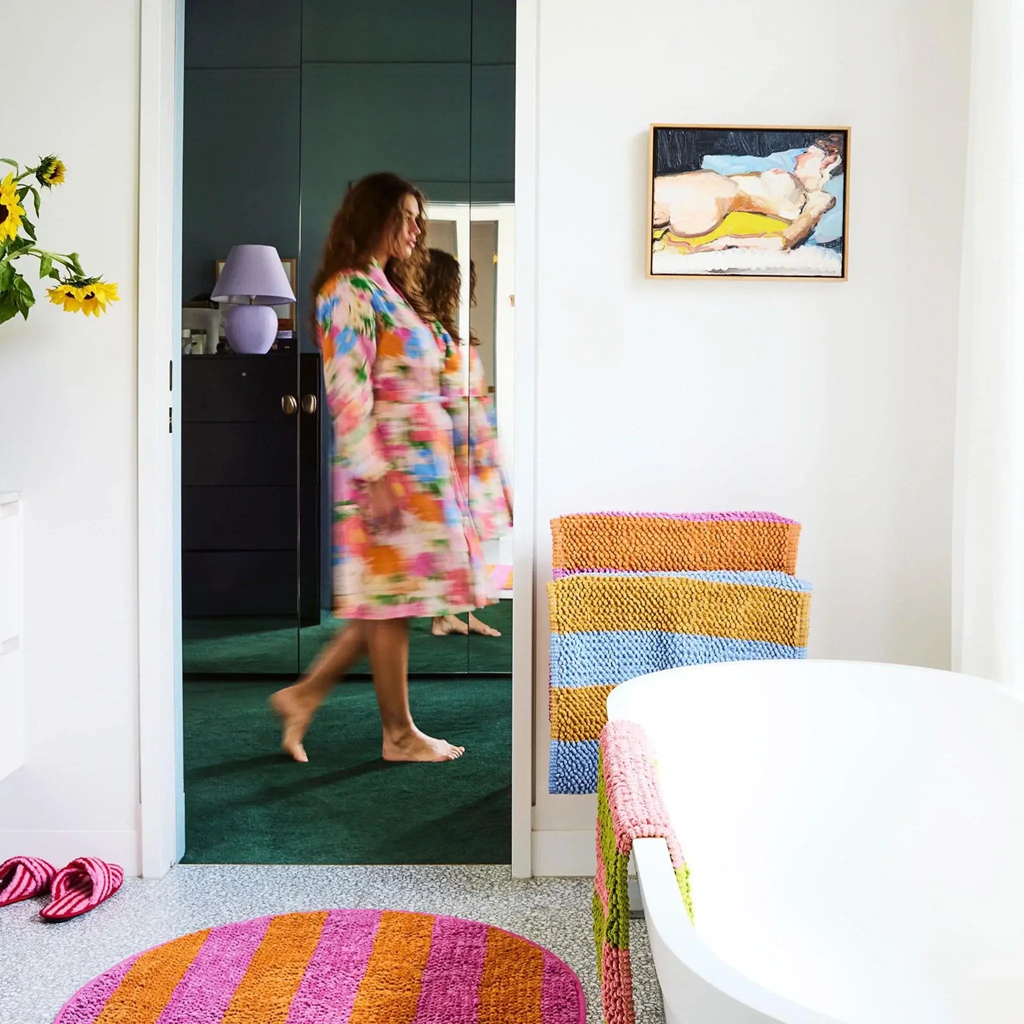 Woman in a colorful robe walking into a bathroom with striped bath mats and a painting on the wall.