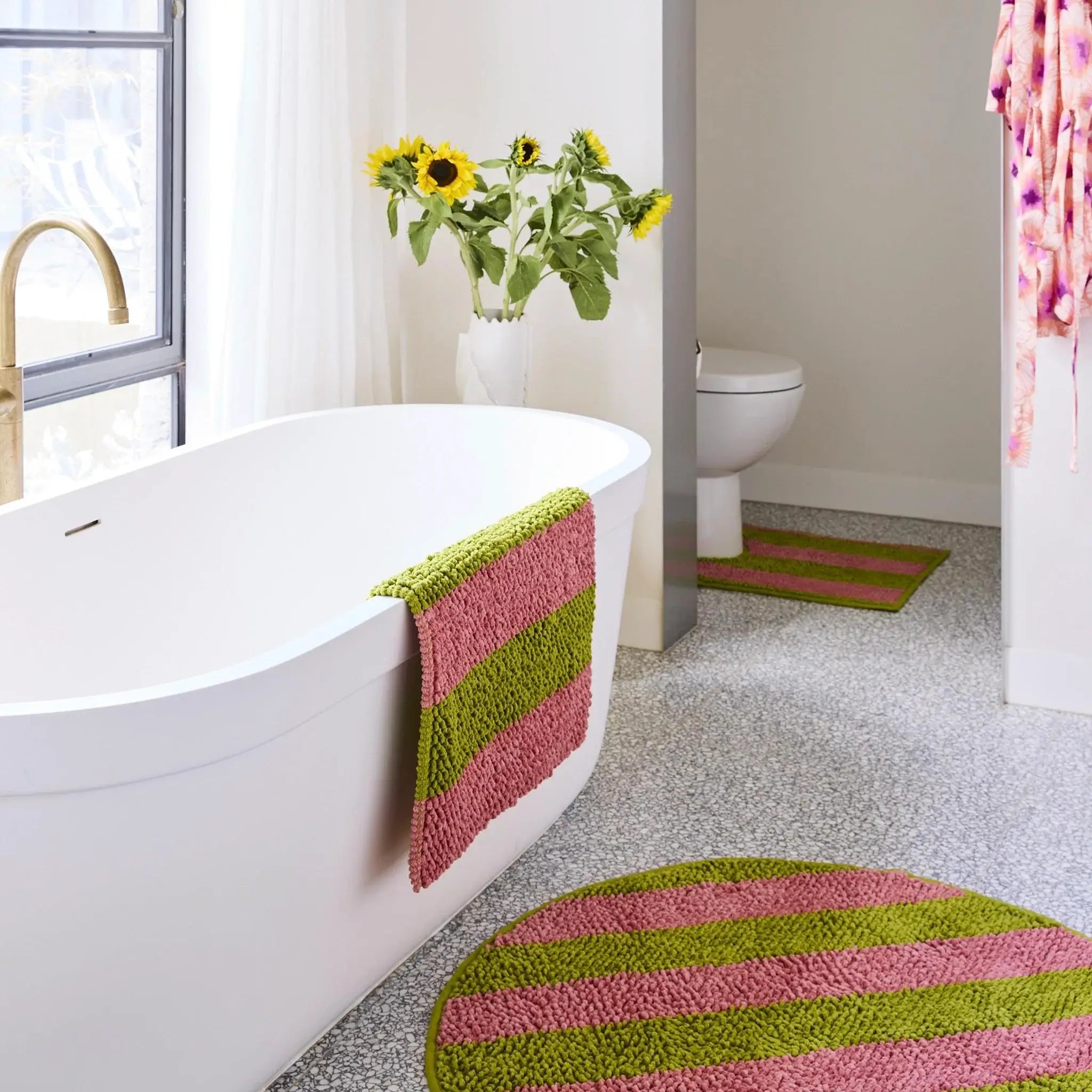 Bathroom with a white bathtub, striped green and pink bath mat, and sunflowers on a windowsill.