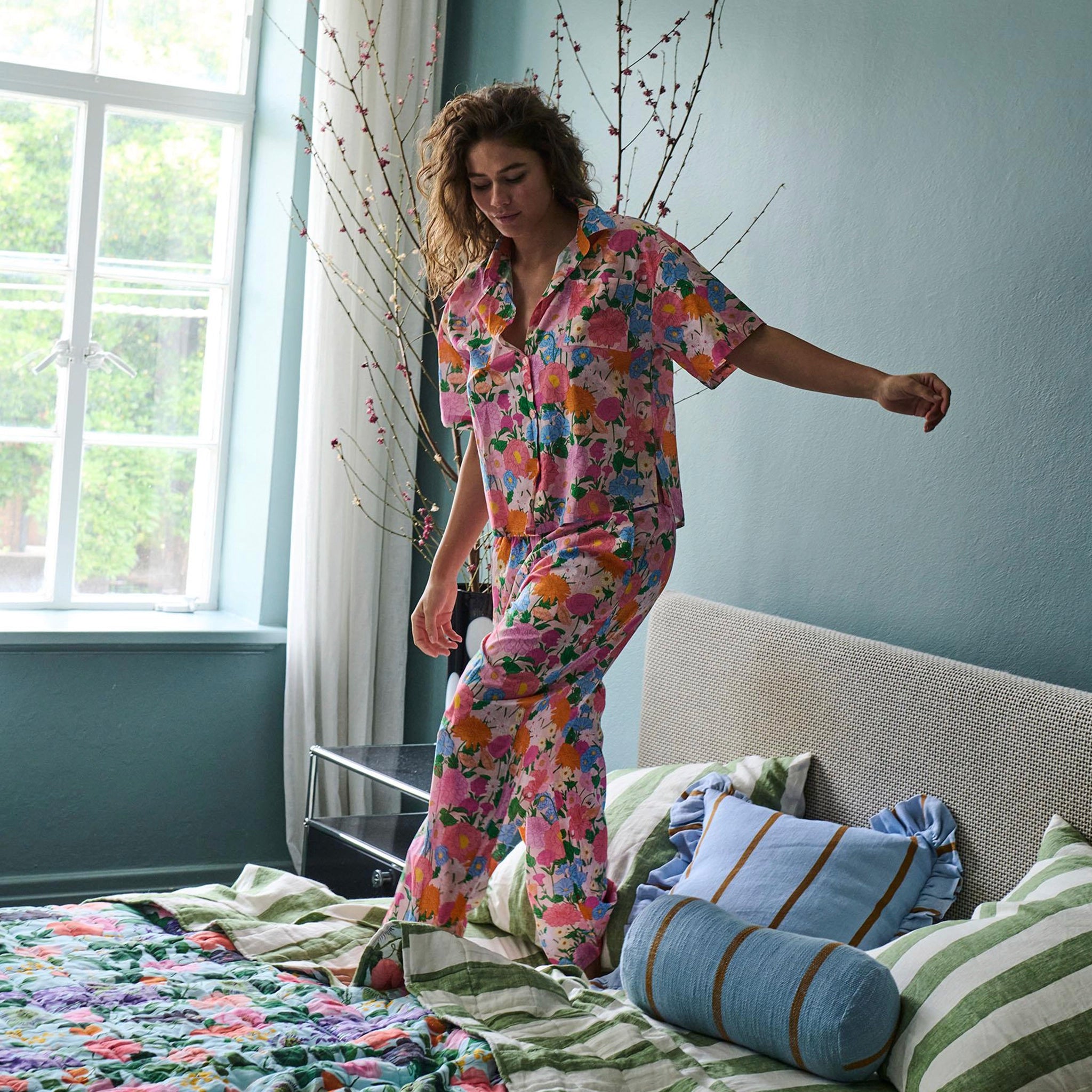 Woman in floral pajama set standing on a bed in a bedroom.
