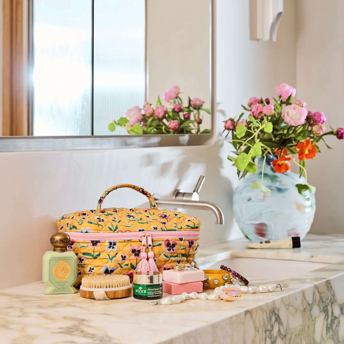 Kip & Co Quilted Toiletry Case - Pretty Pansy on bathroom countertop with various toiletries and flowers.