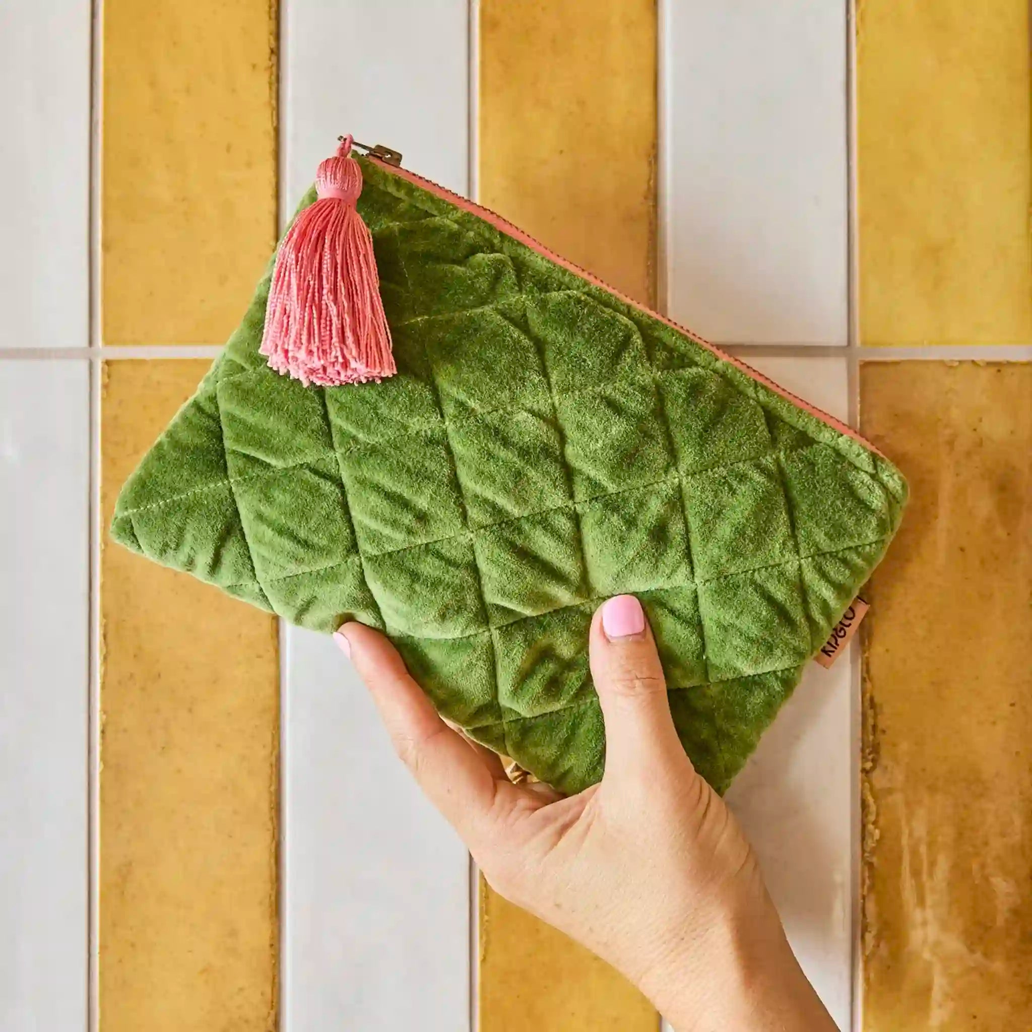 Green textured clutch with a pink tassel held by a hand against a tiled wall background