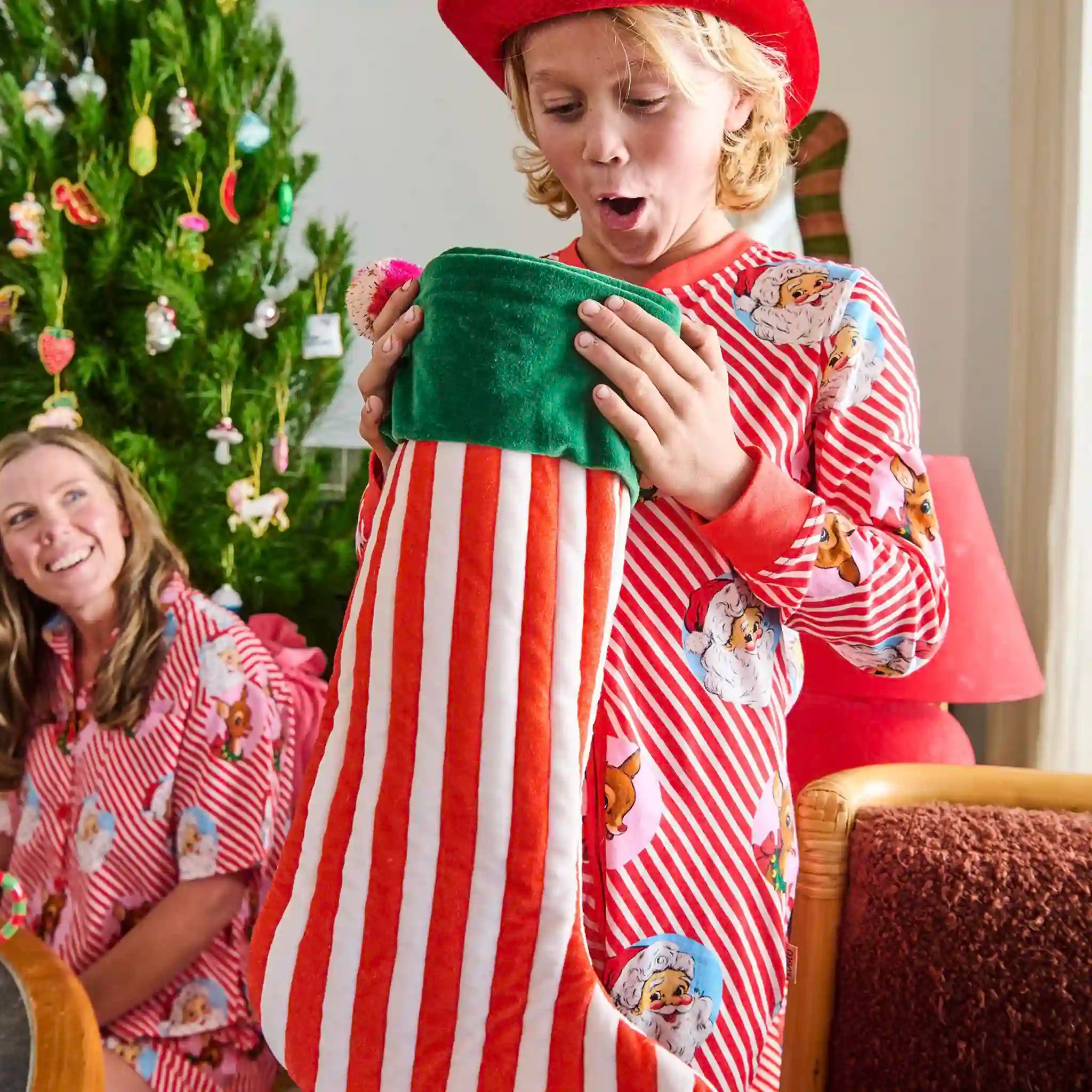 Child in a red and white striped pajama with cartoon characters, holding a green pillow, with a woman in a similar pajama in the background.